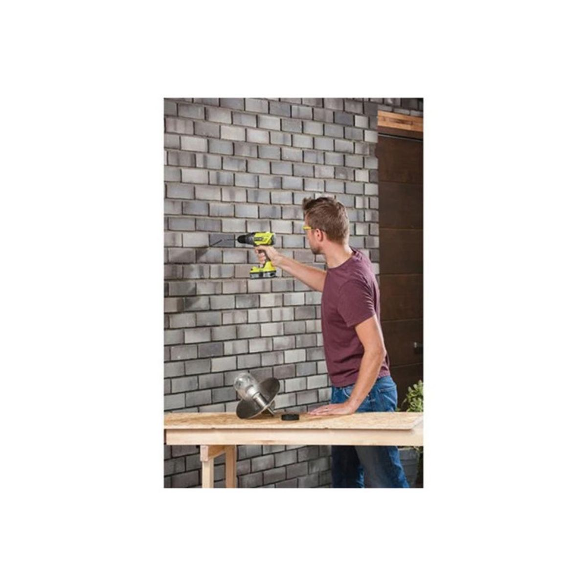 Un uomo con una maglietta bordeaux usa un trapano a batteria su un muro di mattoni grigi.