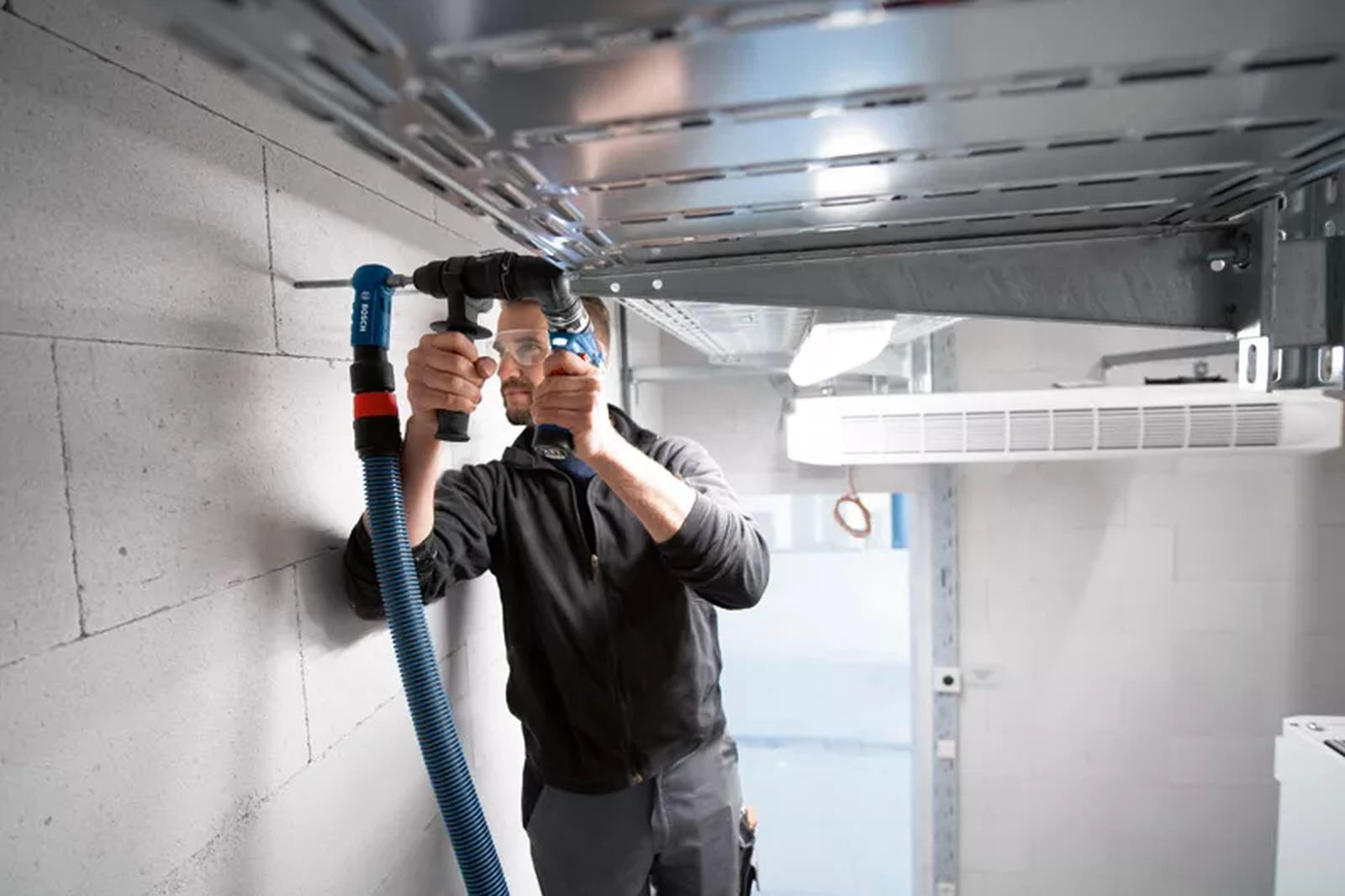 Un lavoratore con dispositivi di sicurezza fora una parete in cemento con un trapano a batteria collegato a un tubo blu.
