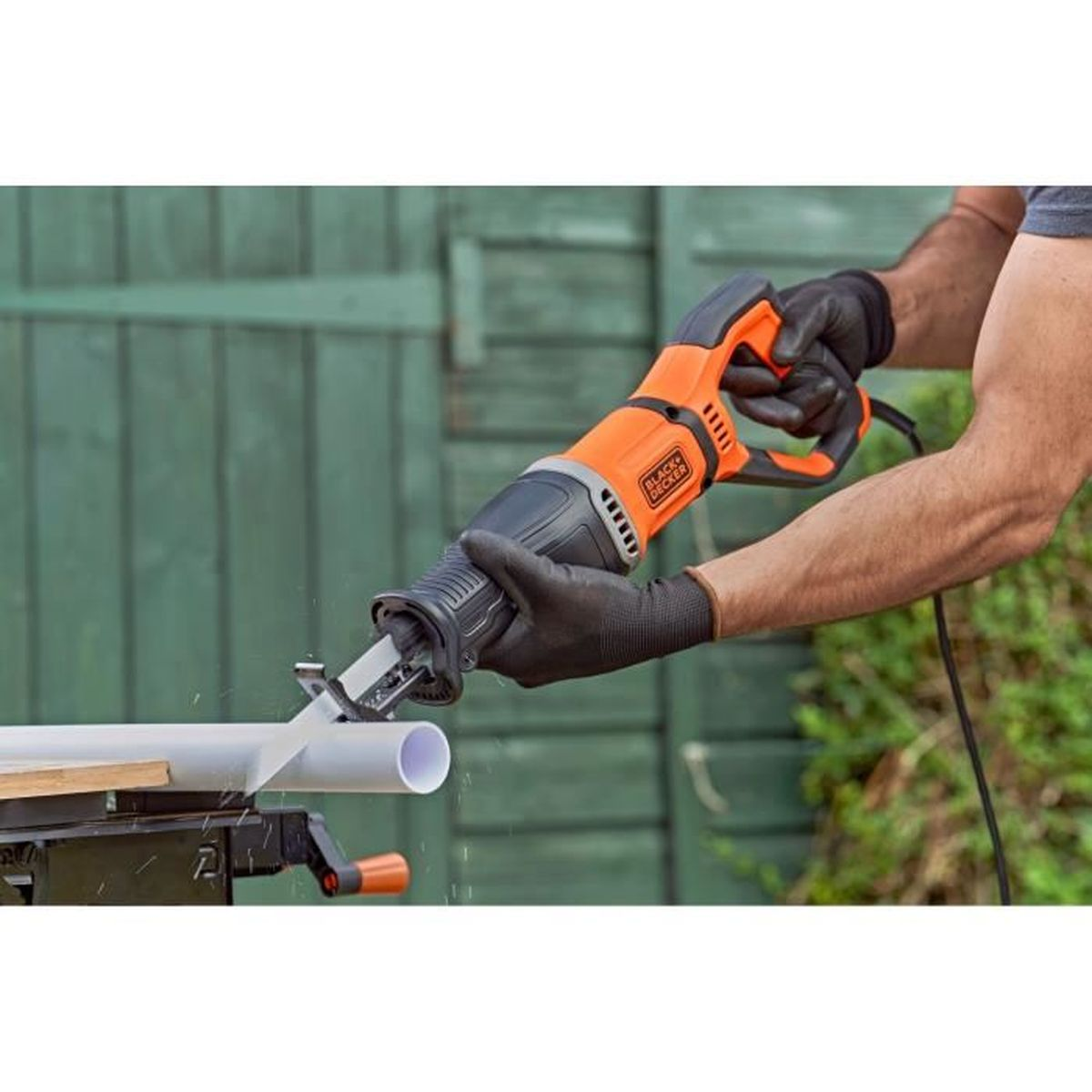 Eine Person mit schwarzen Handschuhen schneidet draußen mit einer orange-schwarzen Säge ein weißes Rohr.