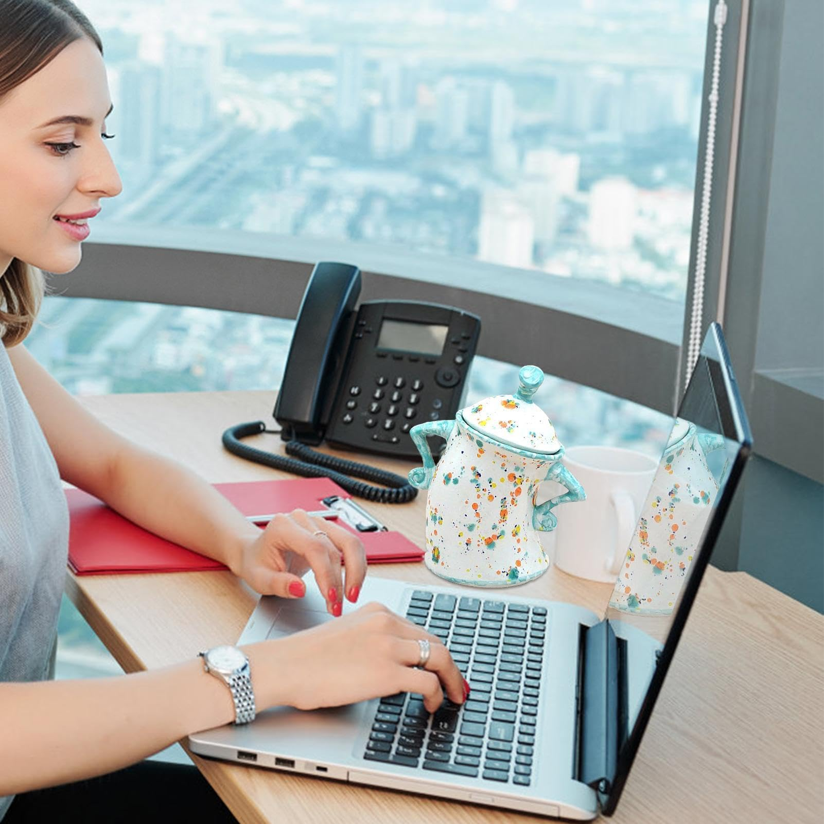 Eine Frau tippt im Büro am Laptop, mit Telefon und bunten Keramikobjekten auf dem Tisch vor einem Stadtblick.