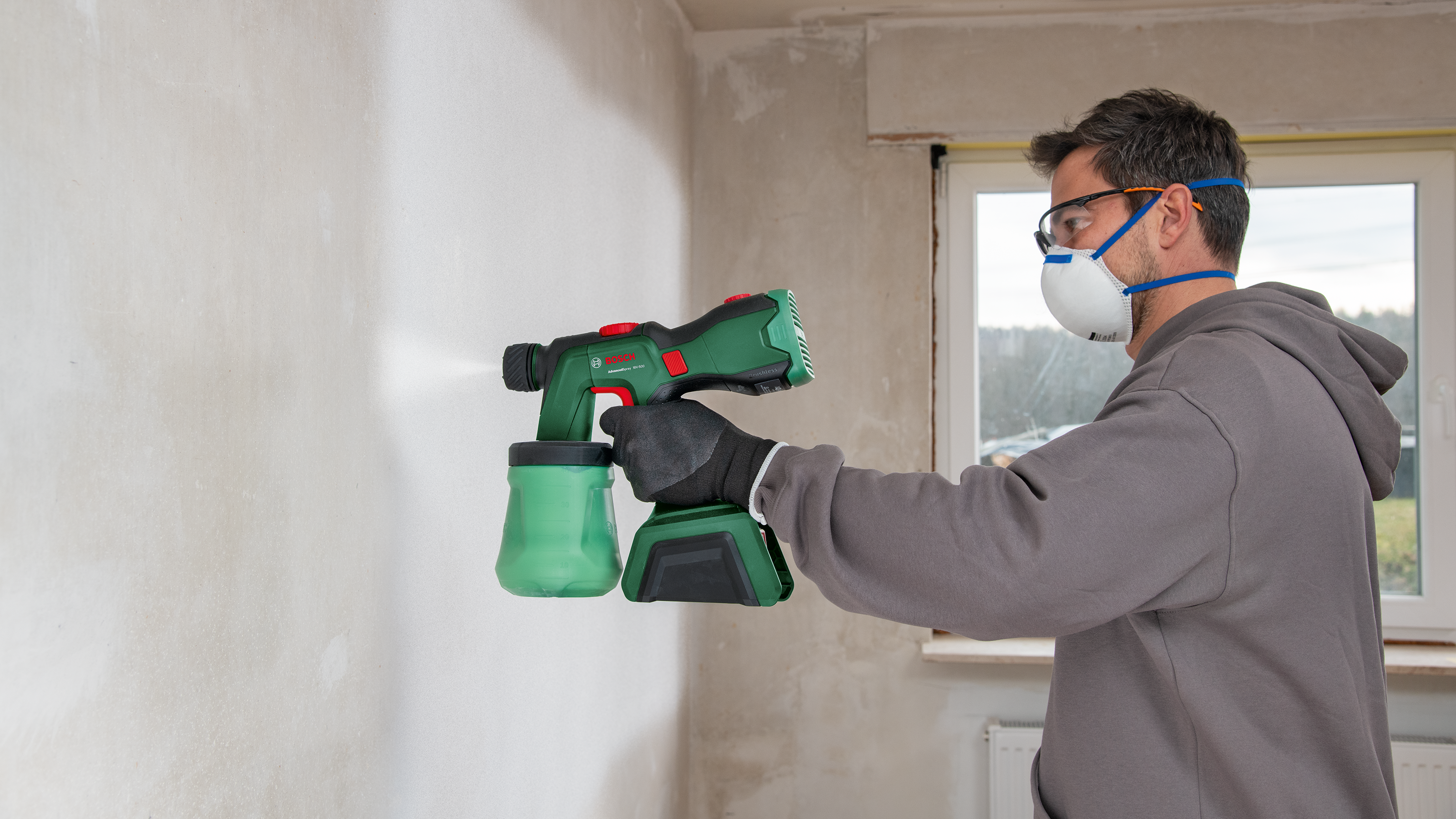 Ein Handwerker mit Schutzbrille und Maske sprüht mit einem grünen Farbsprühgerät Farbe auf eine weiße Wand.