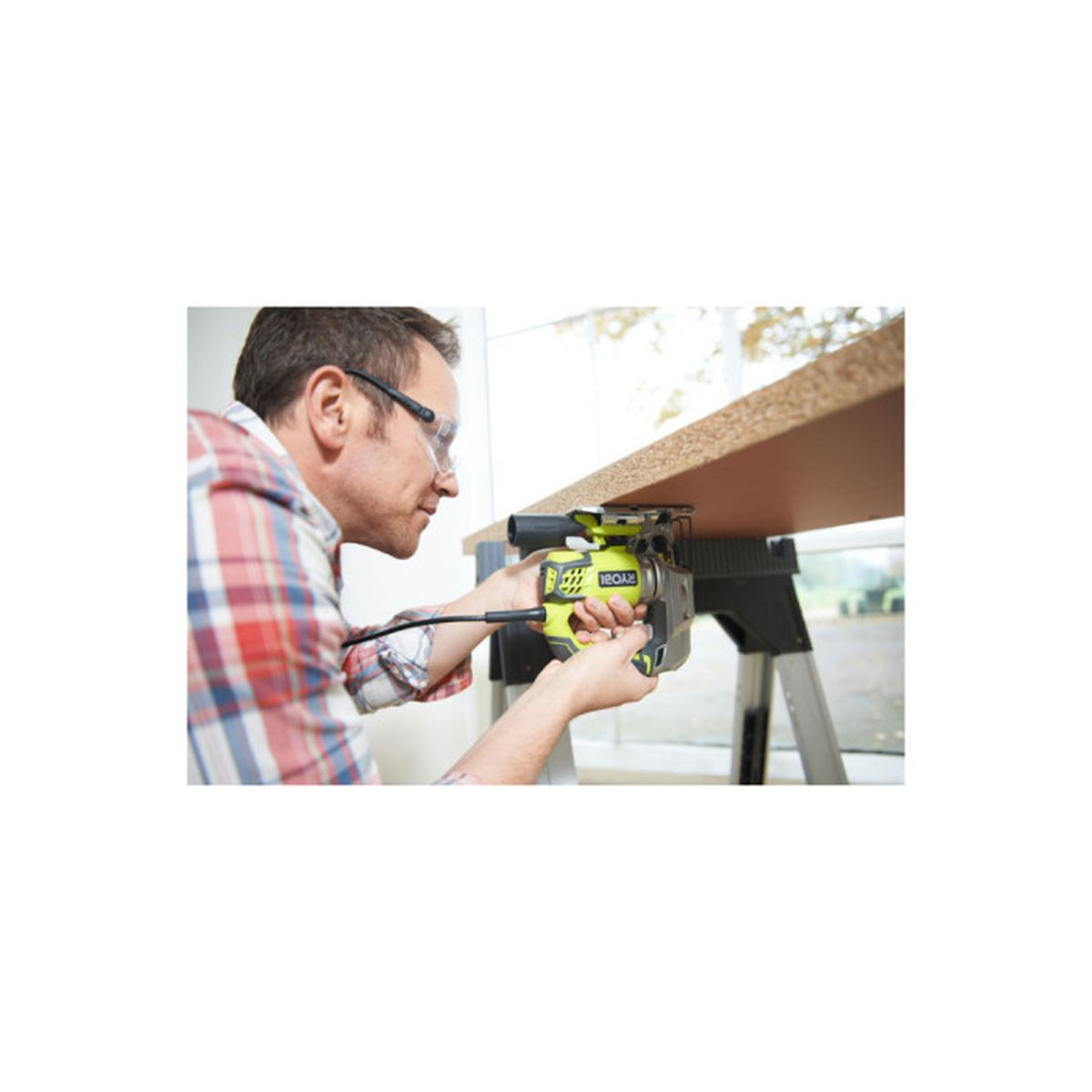 Ein Mann mit Schutzbrille und kariertem Hemd schneidet in einem hellen Raum Holz mit einer Stichsäge.