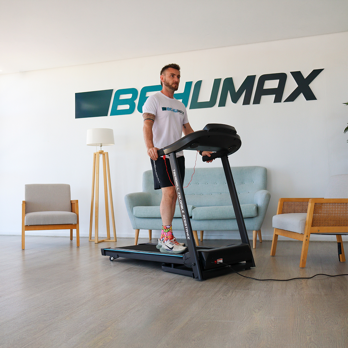 Un hombre con ropa deportiva corre en una cinta de correr negra en una sala luminosa con sillones y logotipo.