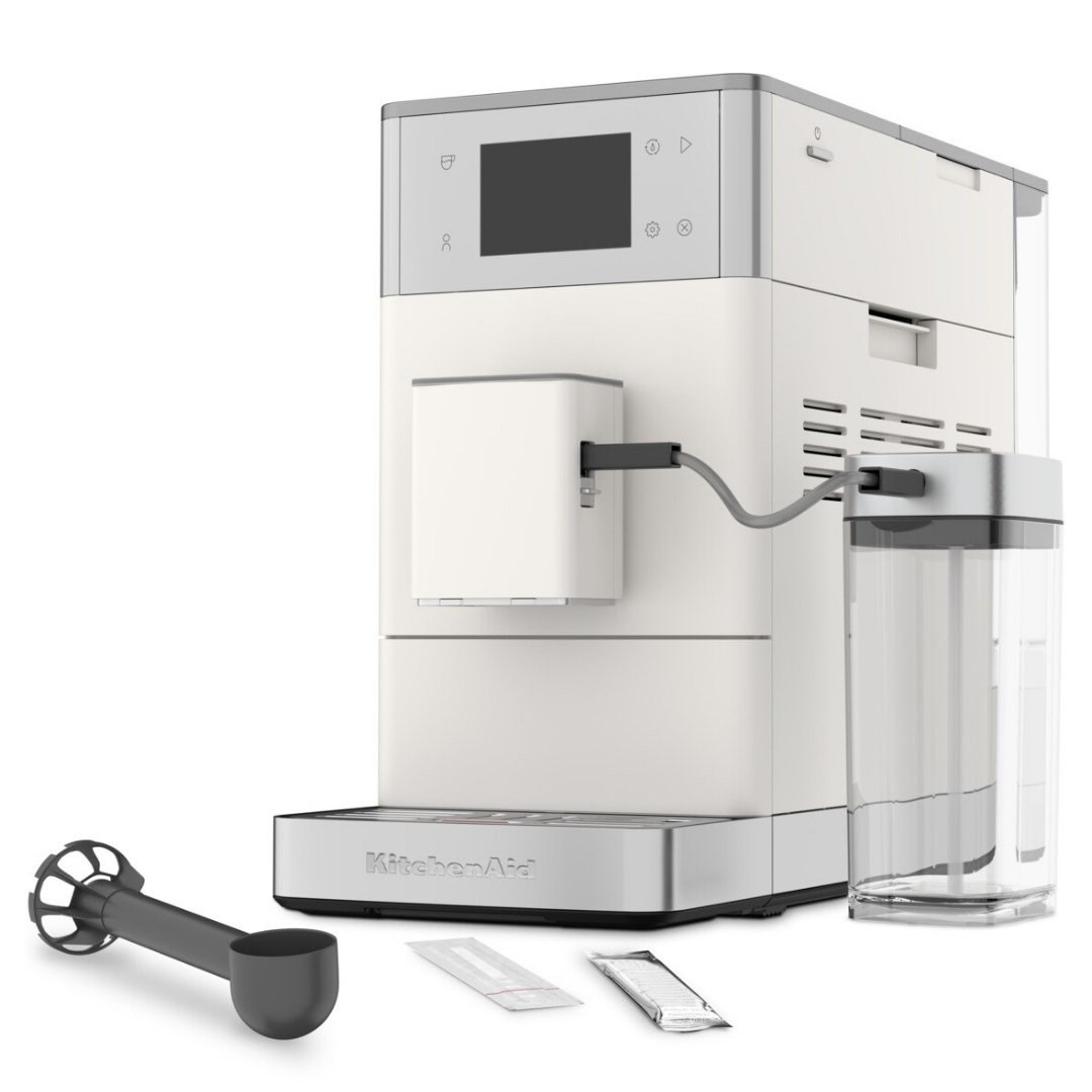 Süt hazneli, kaşıklı ve temizlik poşetli beyaz KitchenAid kahve makinesi.