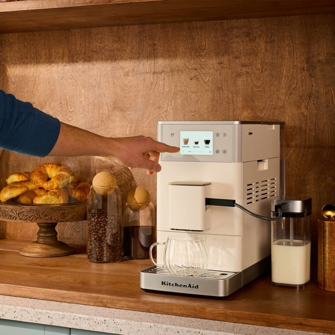 Bir el, ahşap tezgah üzerinde ekranlı beyaz bir KitchenAid kahve makinesini kullanıyor.
