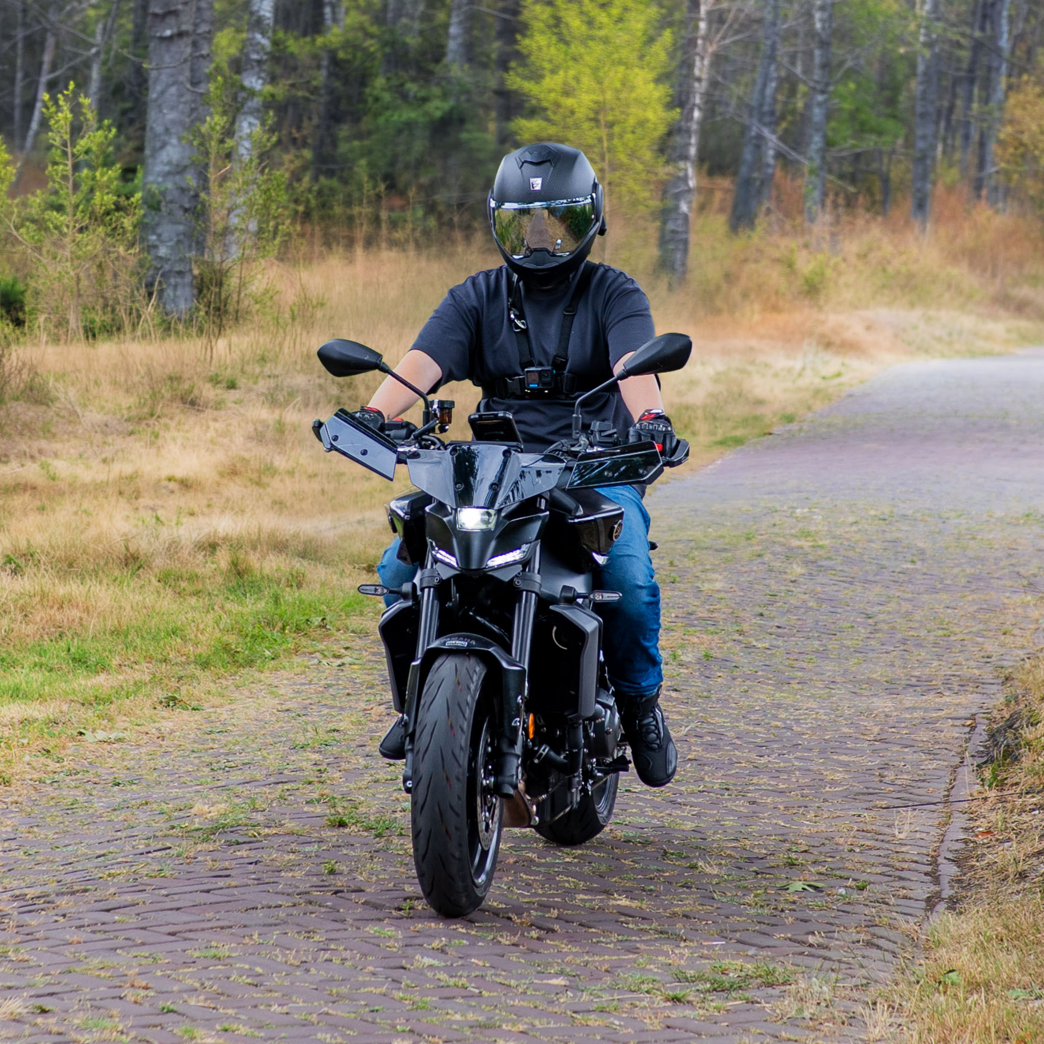 Een motorrijder met een zwarte helm en T-shirt rijdt op een zwarte motor over een verhard pad in een bos.