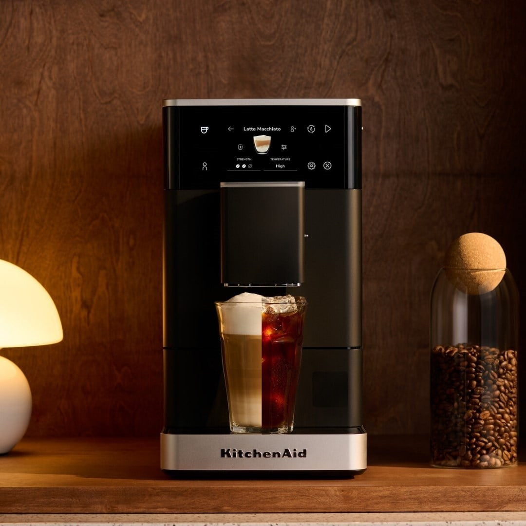 Ahşap tezgah üzerinde siyah ve gümüş renkli KitchenAid kahve makinesi, katmanlı kahve bardağı ve çekirdek kavanozu.
