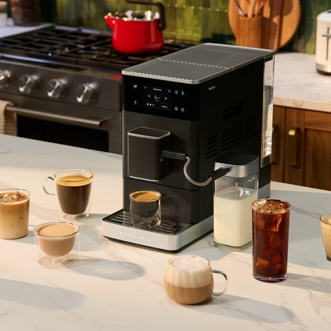Beyaz tezgah üzerinde siyah espresso makinesi, çeşitli kahve fincanları ve bir bardak süt.