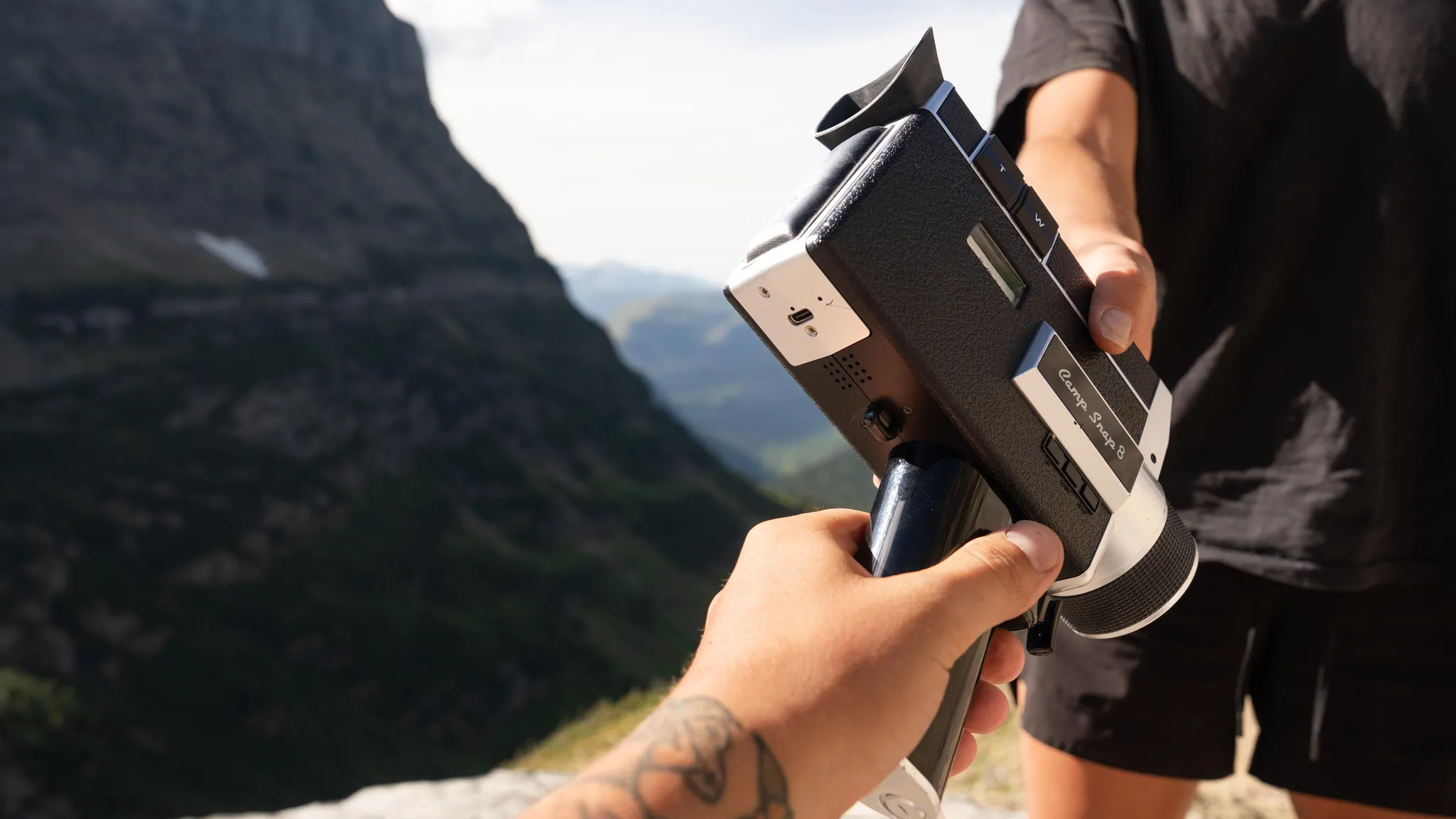Eine Person übergibt eine alte schwarz-weiße Filmkamera im Freien mit Bergen im Hintergrund.