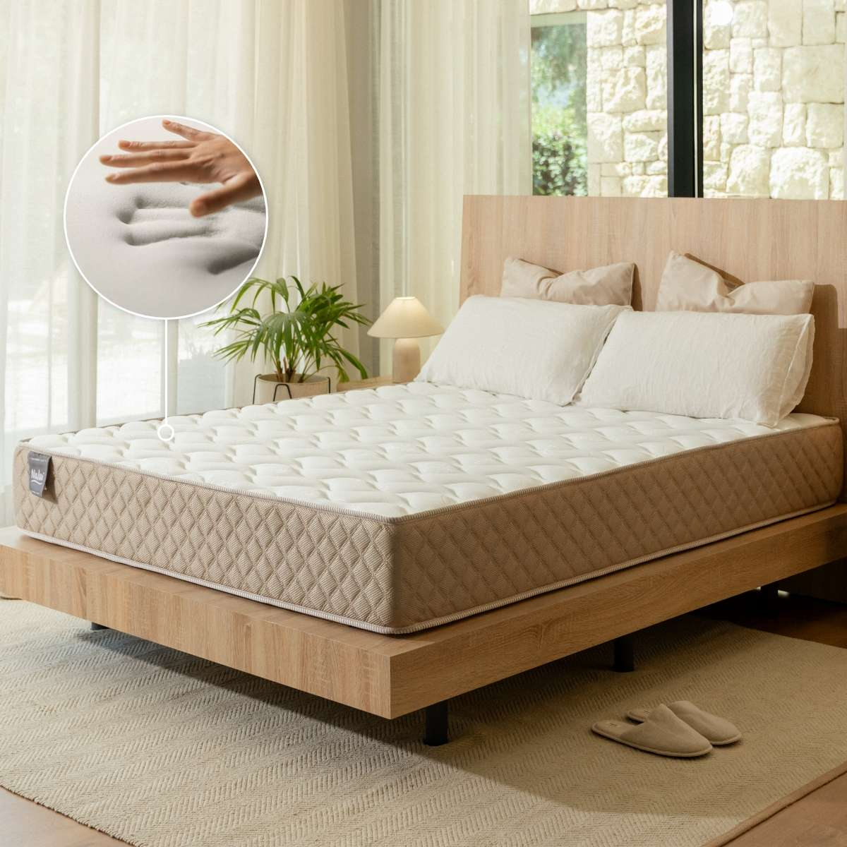 Una cama blanca y marrón con colchón está en una habitación luminosa. Un círculo muestra una mano presionando la espuma.
