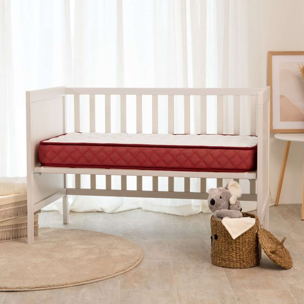 Una cuna de bebé blanca con un colchón rojo y blanco se encuentra en una habitación frente a cortinas blancas.