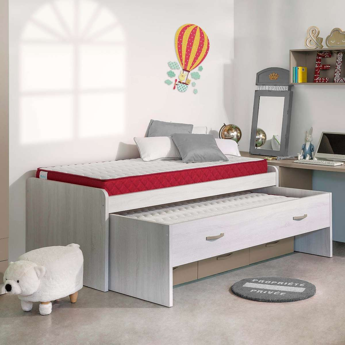 Una cama infantil con nido, colchones rojos y blancos, un taburete de oso y decoración de pared en una habitación luminosa.