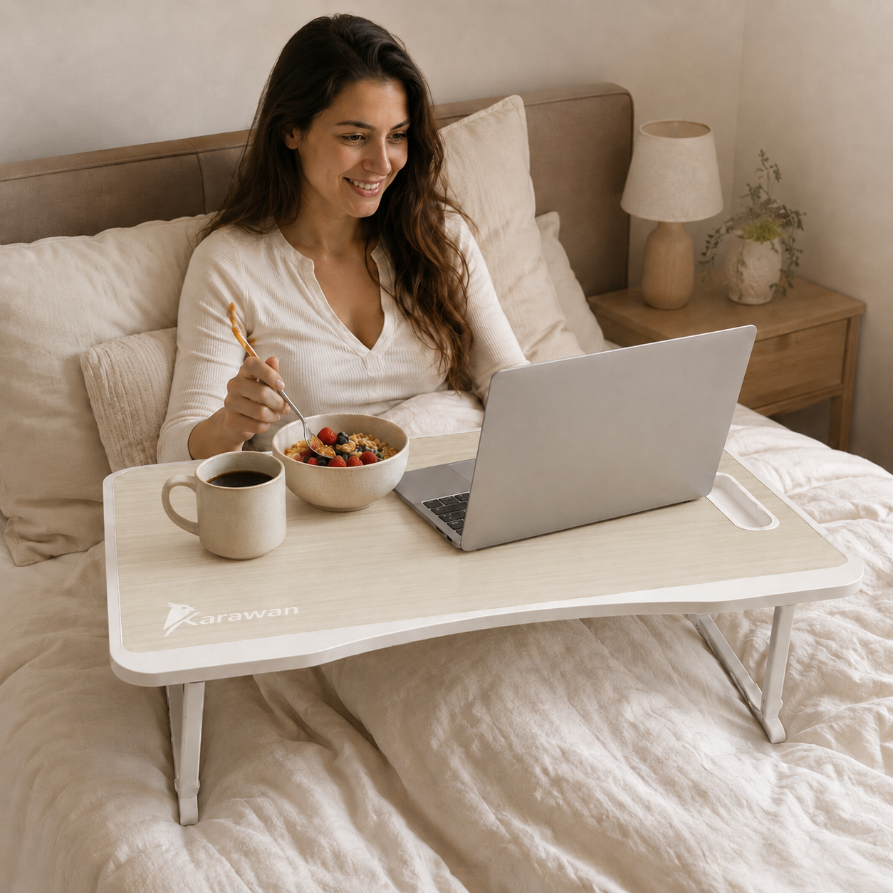 Una mujer sonriente en la cama con portátil, come fruta y bebe de una taza en una mesa auxiliar.