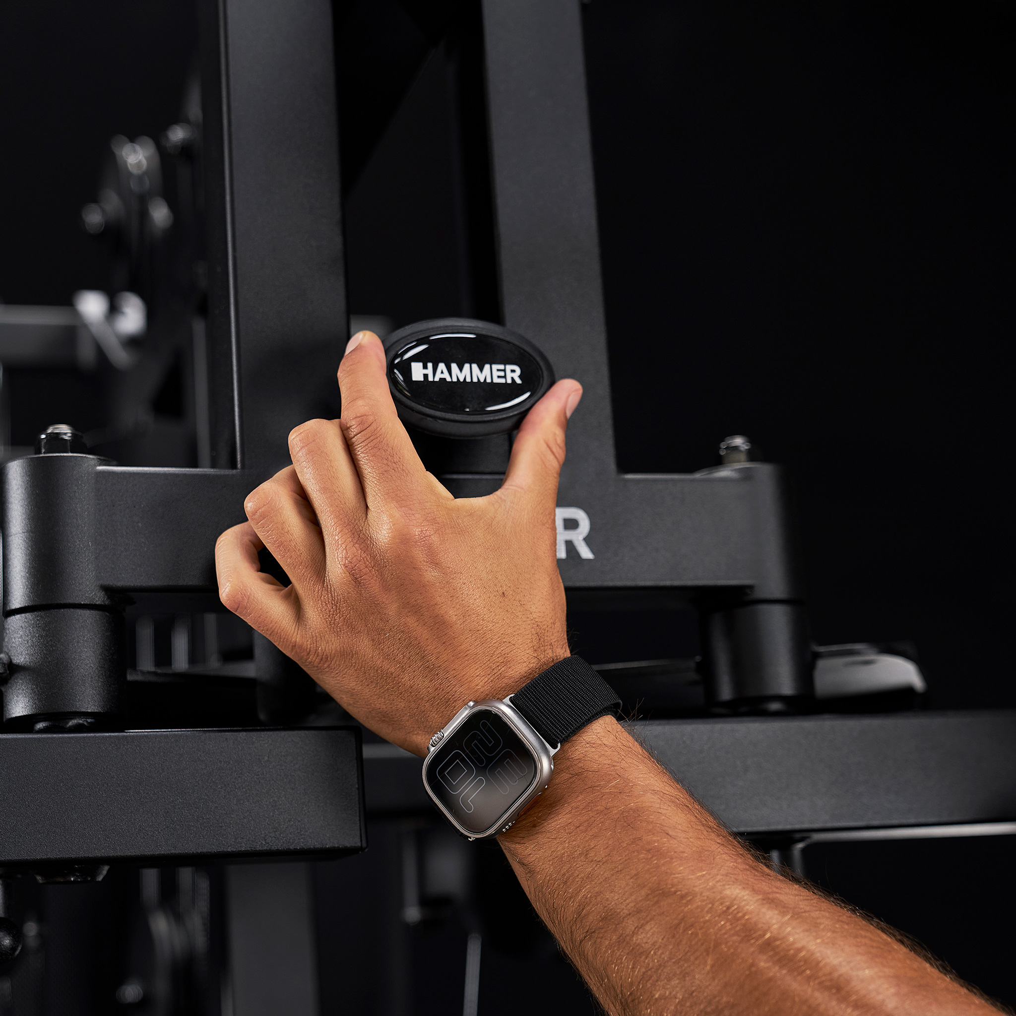 Eine Hand mit Smartwatch verstellt einen schwarzen Hammer-Fitnessgeräteknopf vor dunklem Hintergrund.