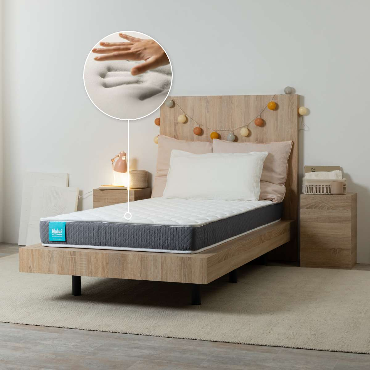 Una cama de madera con colchón gris en un dormitorio, con un acercamiento circular de manos presionando espuma viscoelástica.