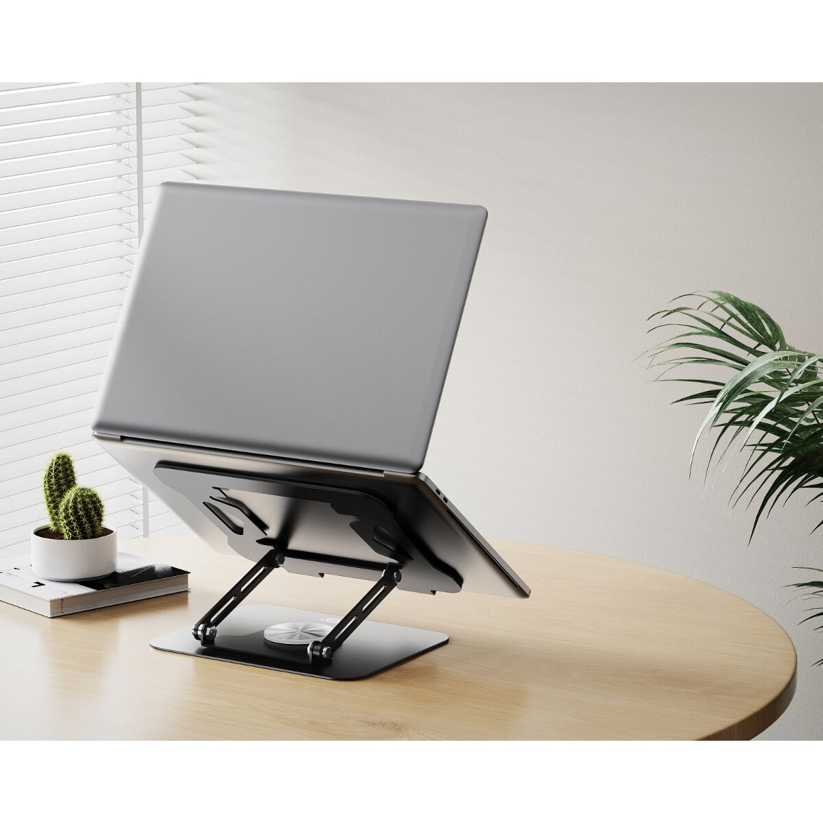 Ein silberner Laptop auf einem schwarzen Ständer auf einem Holztisch, neben einer kleinen Topfpflanze und Jalousien.