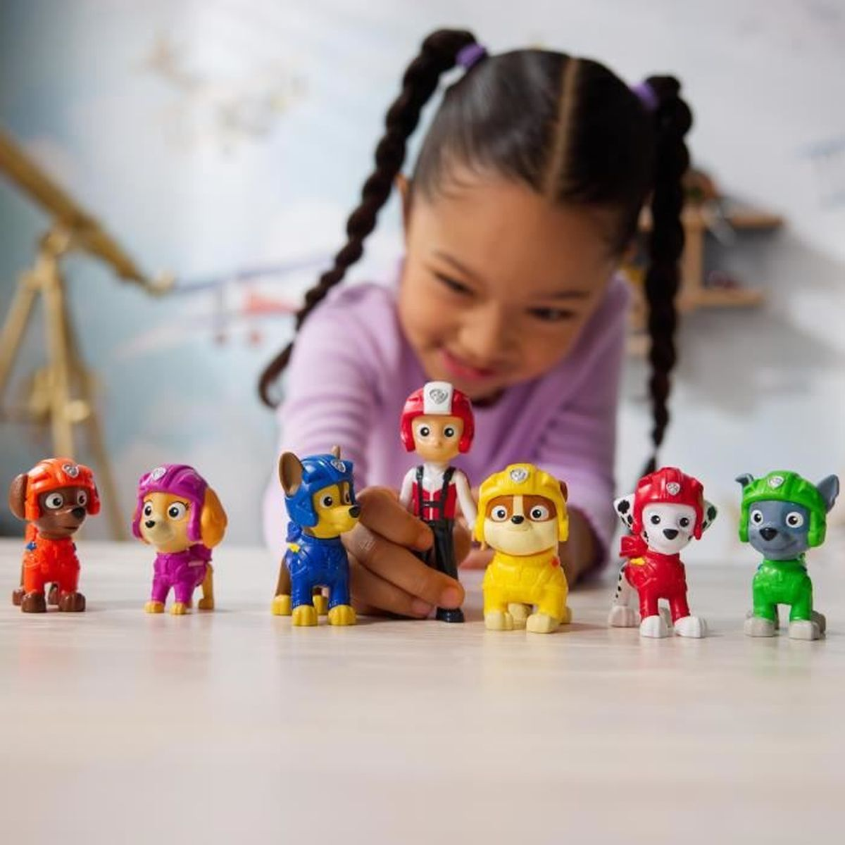 Ein lächelndes Mädchen spielt mit Paw Patrol Figuren auf einem Tisch.