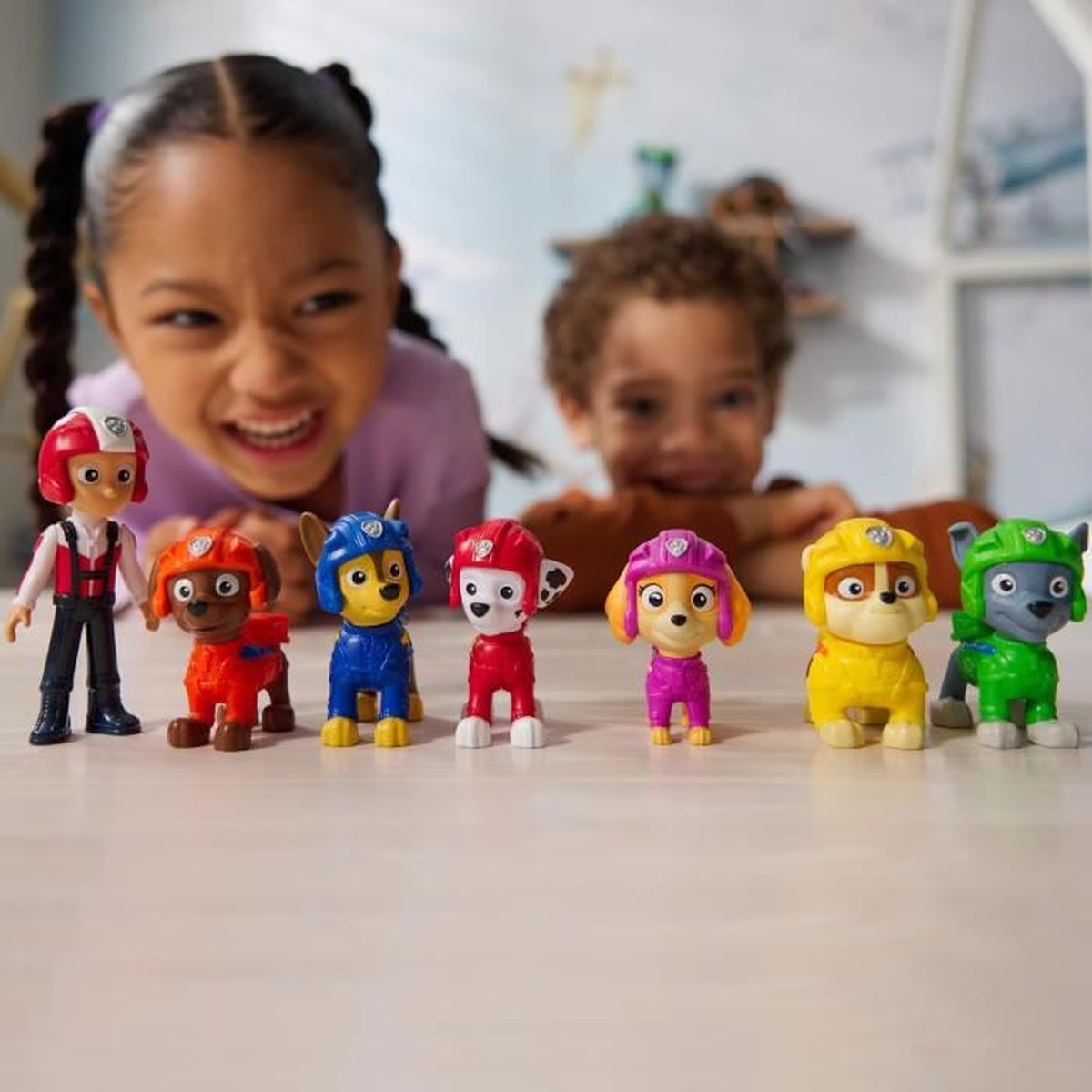 Zwei glückliche Kinder spielen mit Paw-Patrol-Figuren, die in einer Reihe auf dem Tisch stehen.
