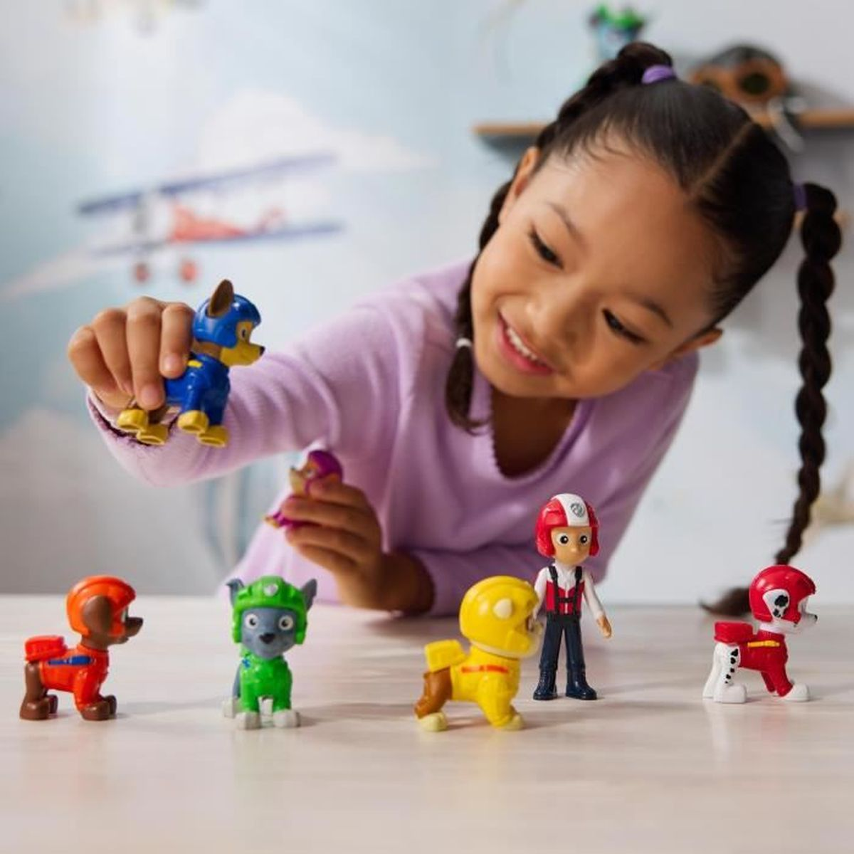 Ein junges Mädchen in einem lila Pullover spielt mit Paw Patrol Figuren auf einem Tisch.