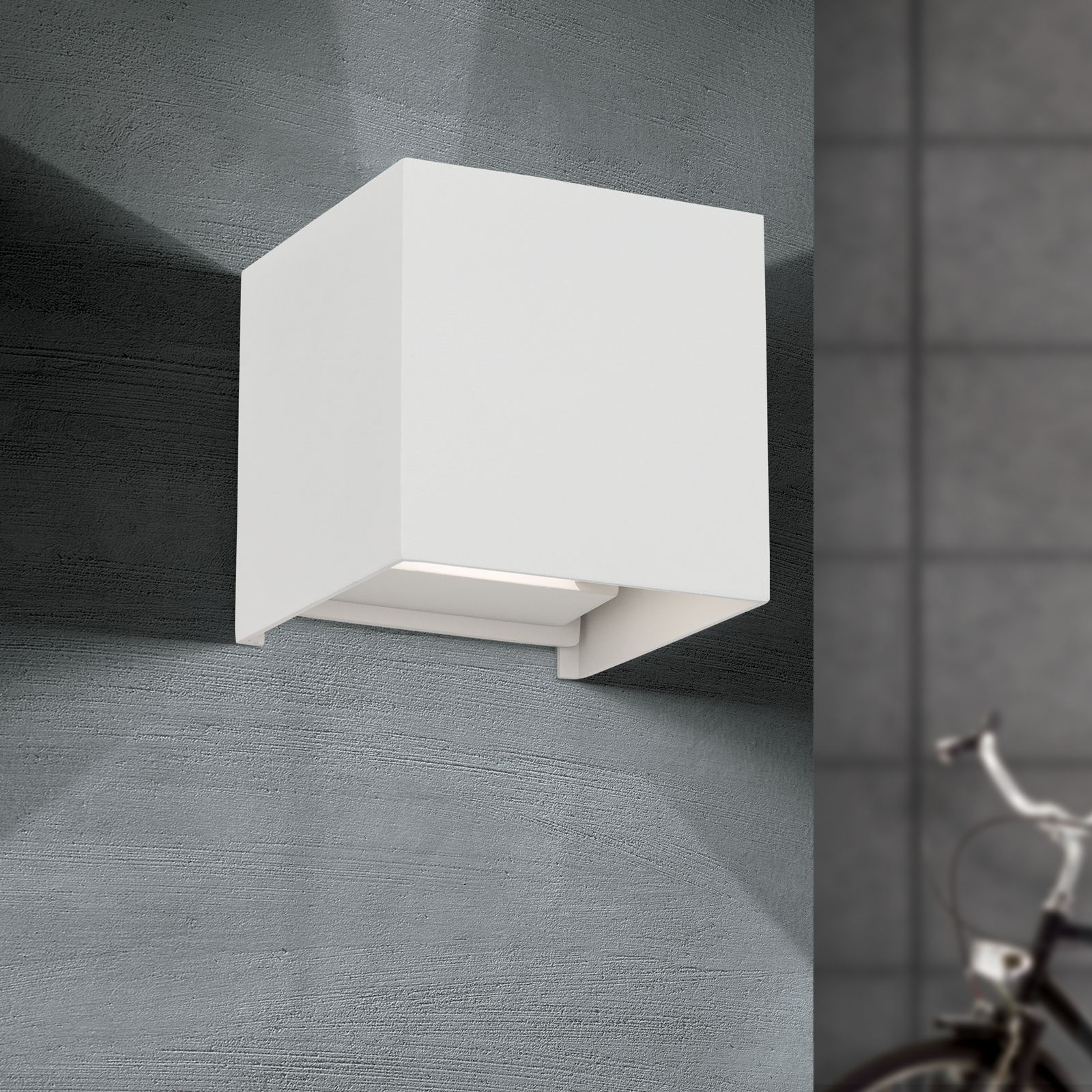 Eine weiße quadratische Wandleuchte an einer grauen strukturierten Wand, mit einer verschwommenen Fahrrad im Hintergrund.