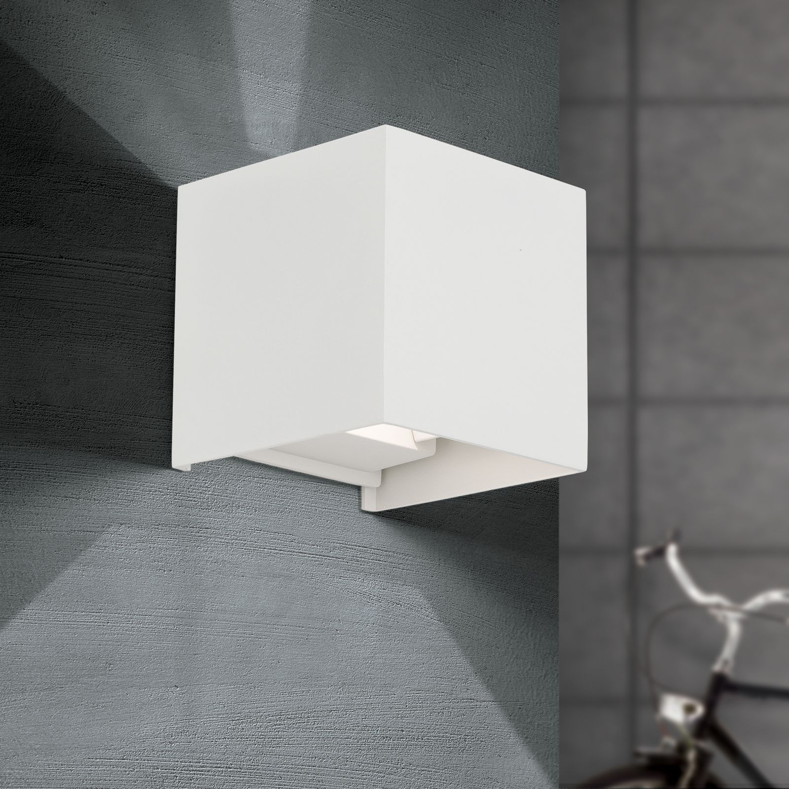 Eine weiße, quaderförmige Wandleuchte an einer dunklen Putzwand wirft Licht nach oben und unten.