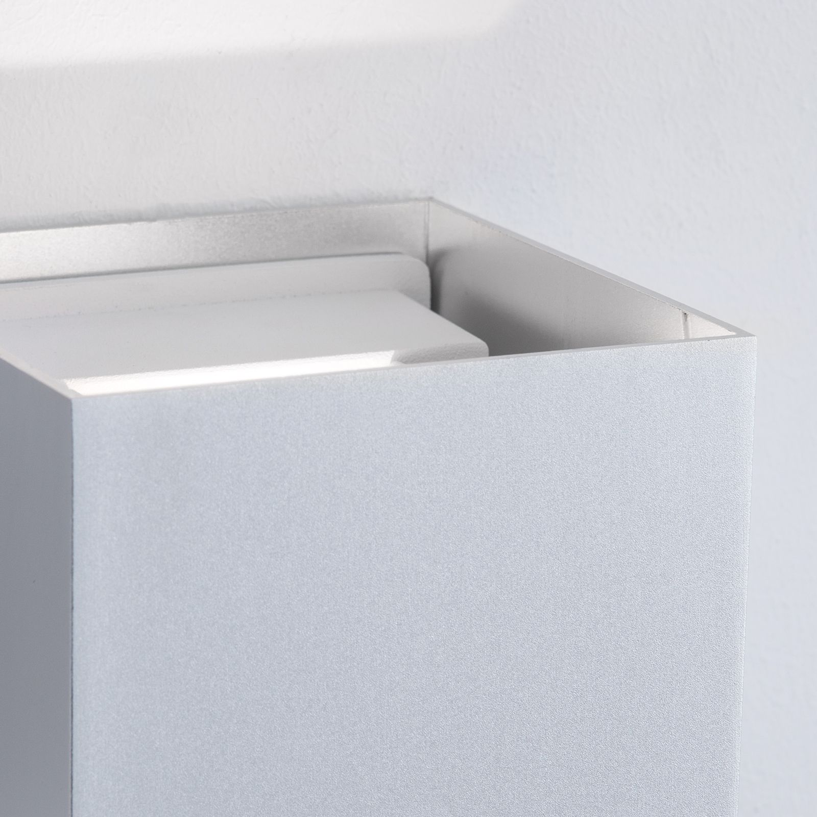 Eine moderne silberne kubische Wandleuchte strahlt sanftes Licht gegen eine weiße Wand aus.
