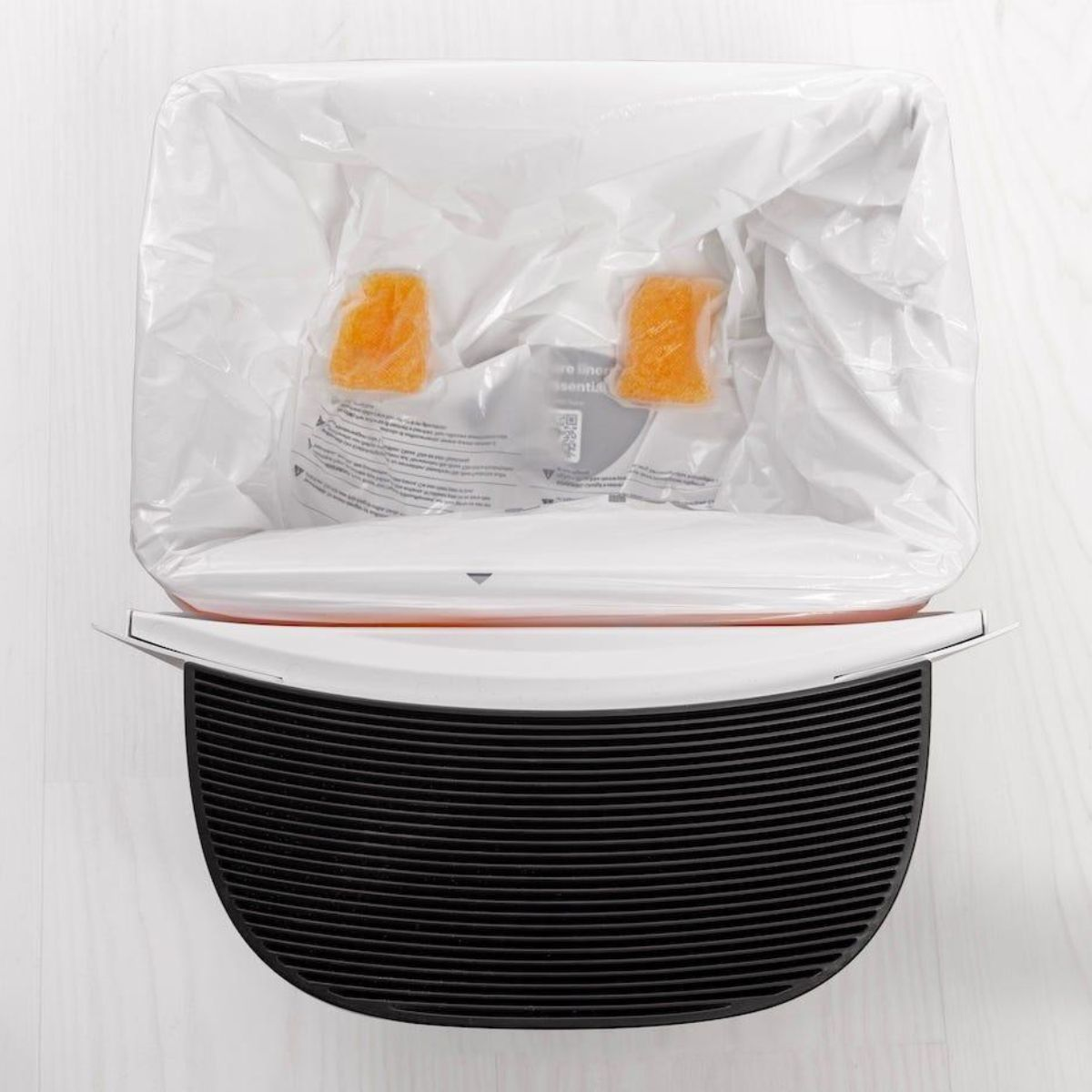 Vista superior de un basurero blanco abierto con una bolsa de plástico blanca y dos recargas naranjas.