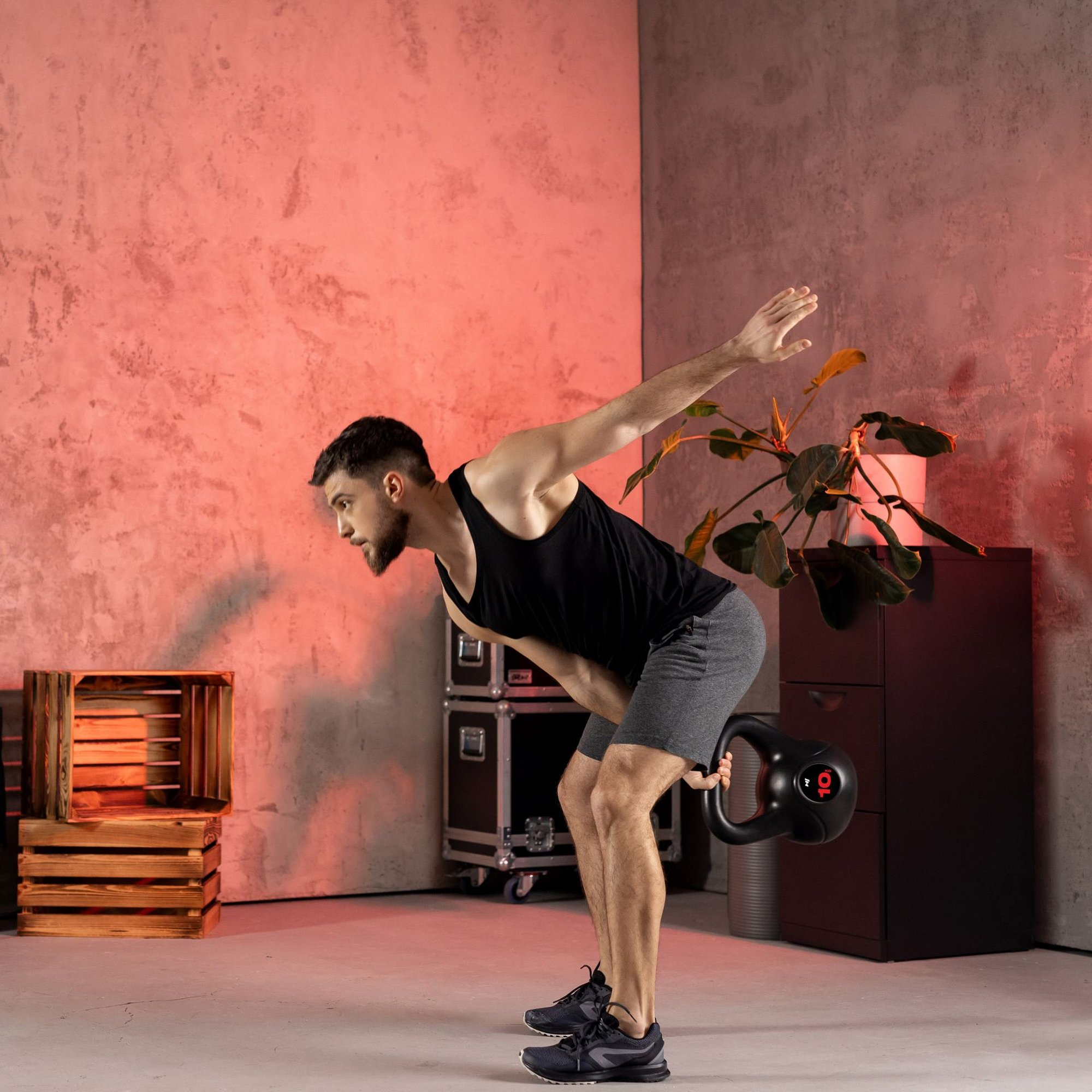 Ein Mann im Sportoutfit hebt eine Kettlebell in einem Raum mit roter Wand und Holzkisten.