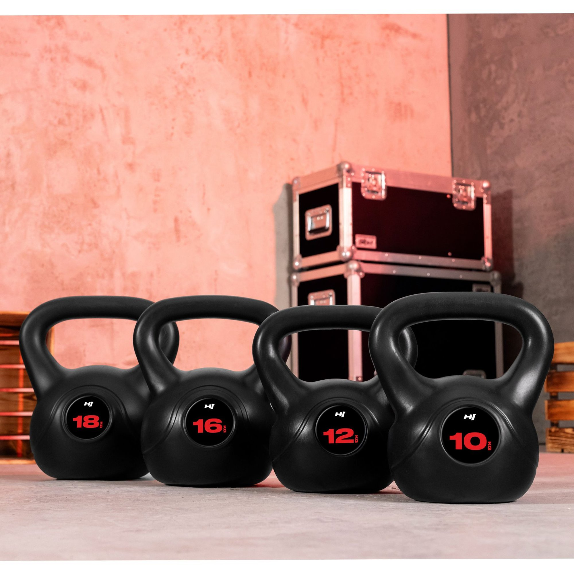 Vier schwarze Kettlebells mit Gewichtsangaben in Rot (18, 16, 12, 10 kg) stehen nebeneinander vor einer rosafarbenen Wand.