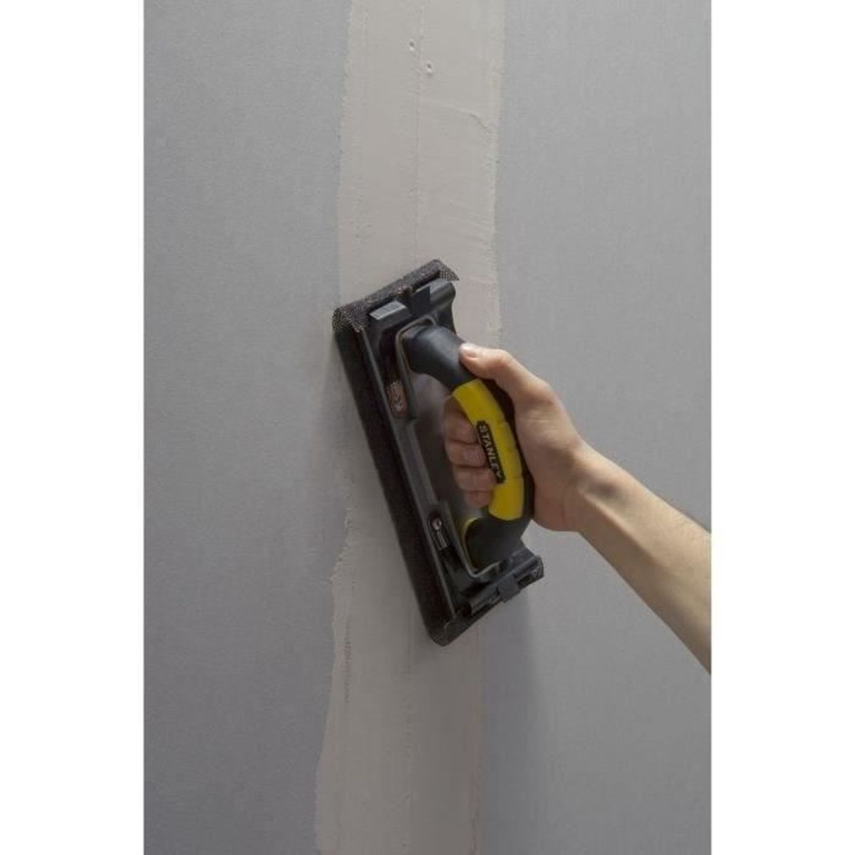 Eine Hand benutzt einen Handschleifer mit gelbem Griff, um eine Trockenbauwand zu schleifen.
