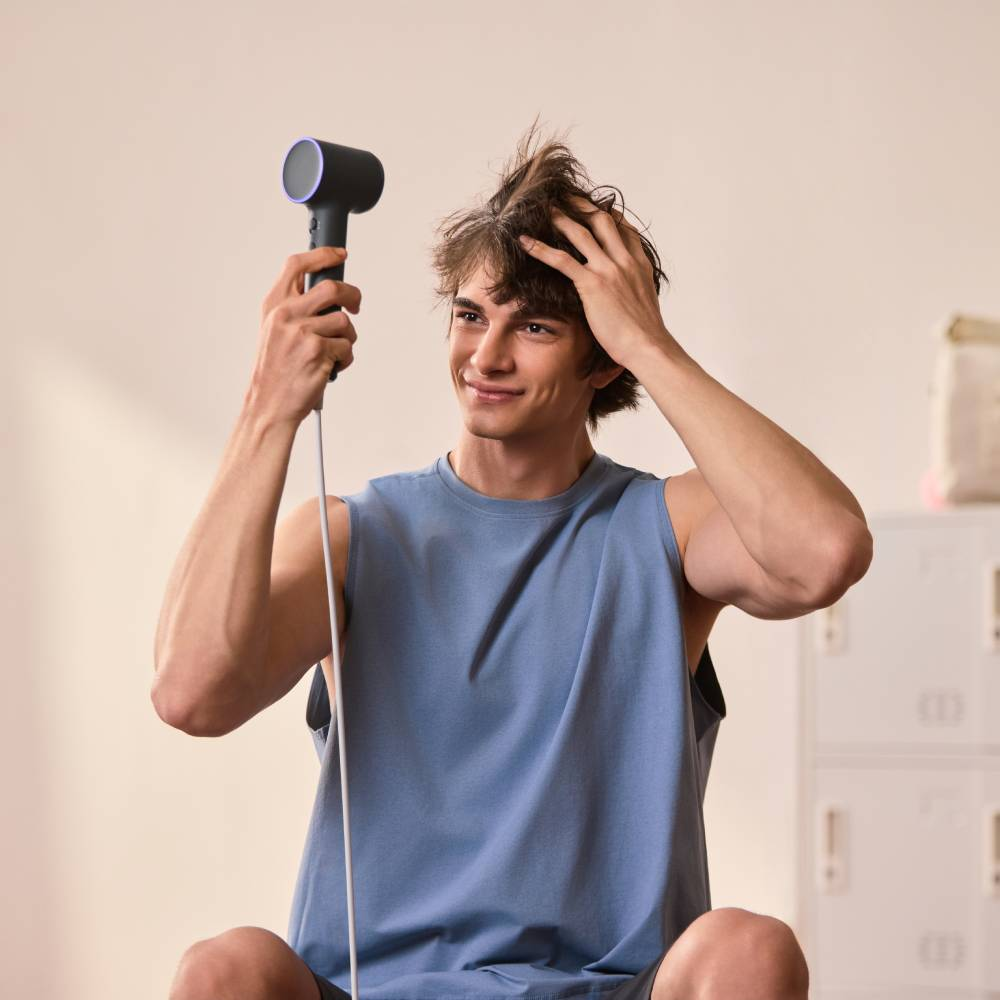 Ein junger Mann im blauen Tanktop hält einen schwarzen Haartrockner und stylt sein Haare in einem hellen Raum.