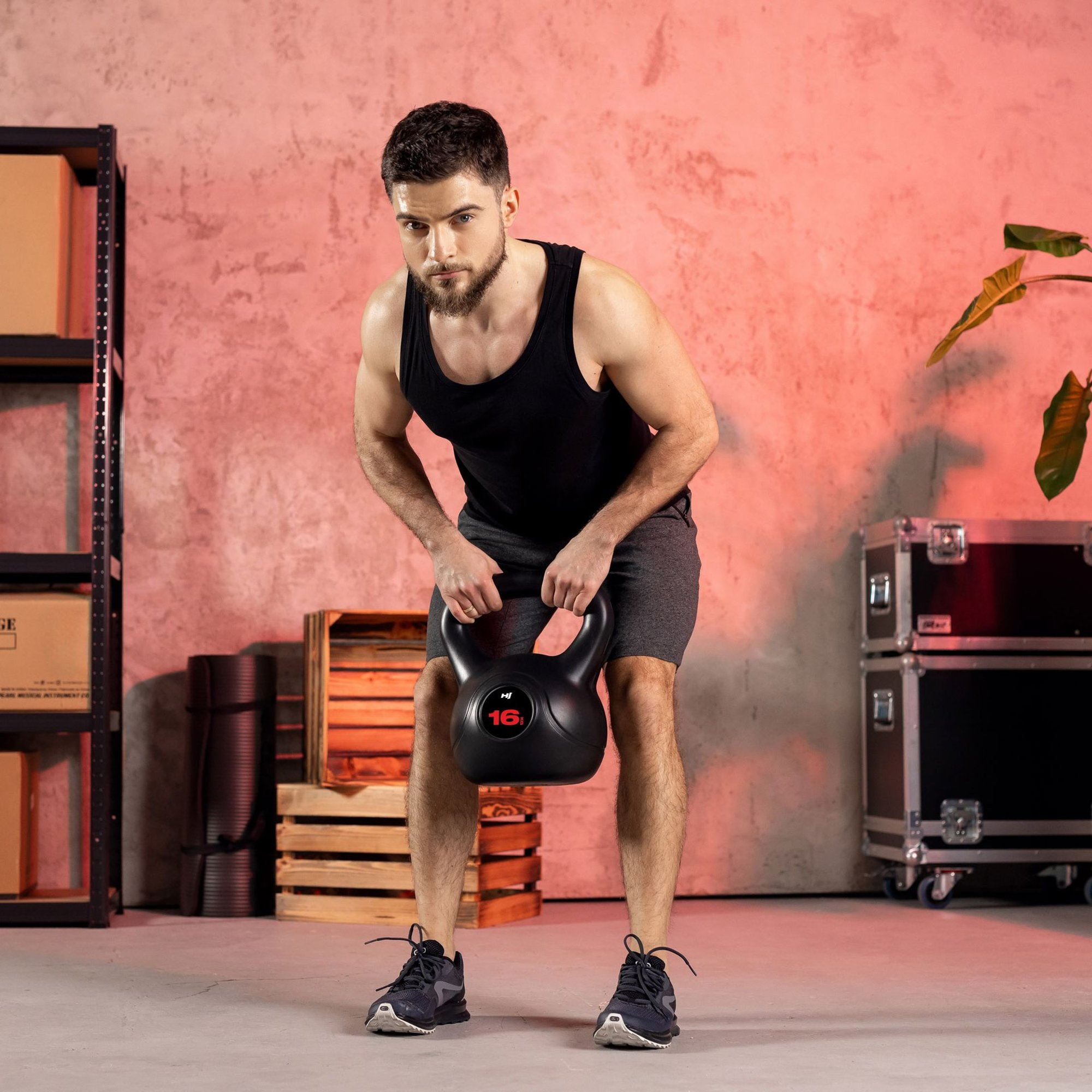 Ein Mann mit Bart in Sportkleidung hebt eine schwere Kettlebell in einem Raum mit roter Wand und Kisten.