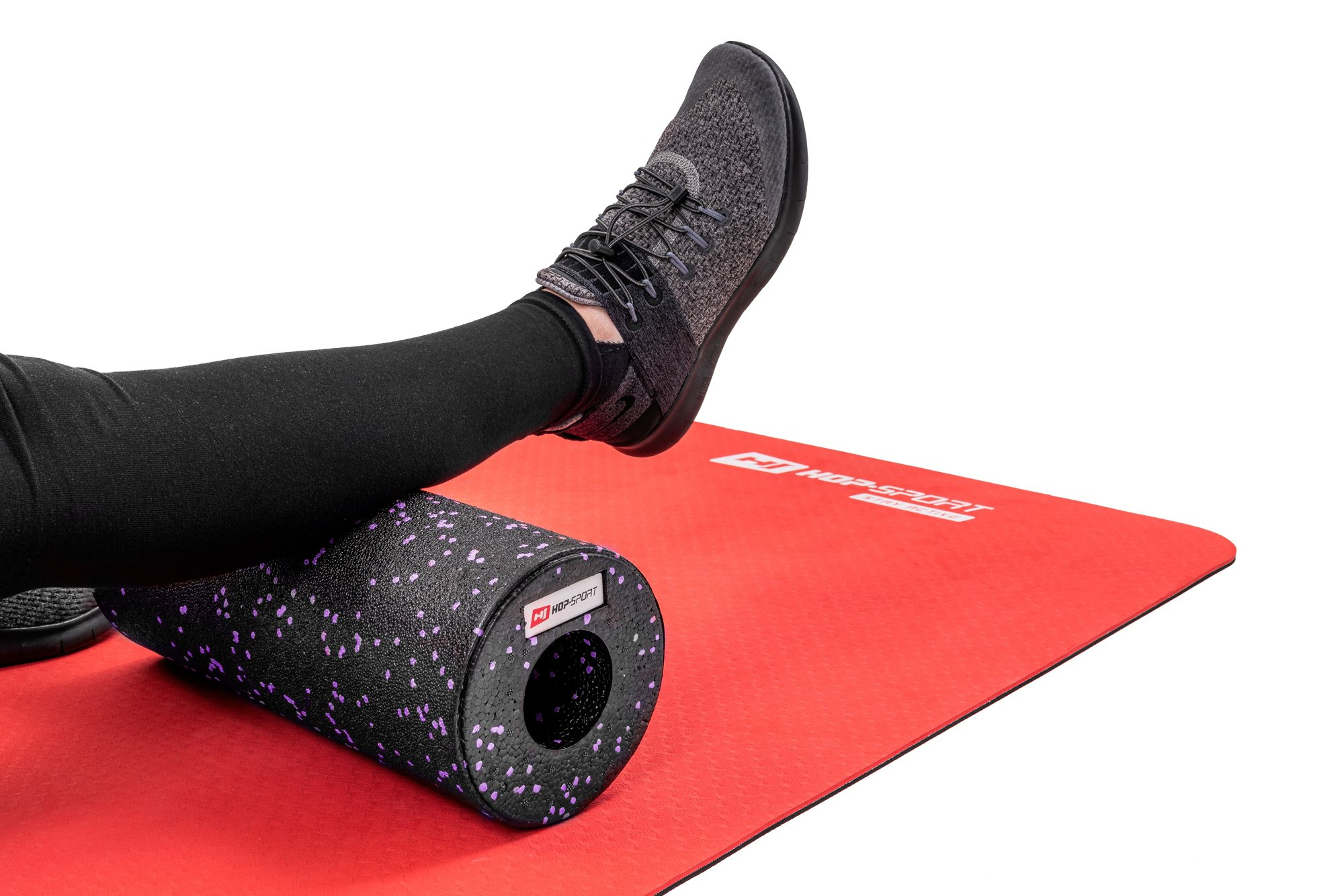 Ein Bein in schwarzer Leggings und grauem Schuh ruht auf einer schwarz-lila Faszienrolle auf roter Matte.