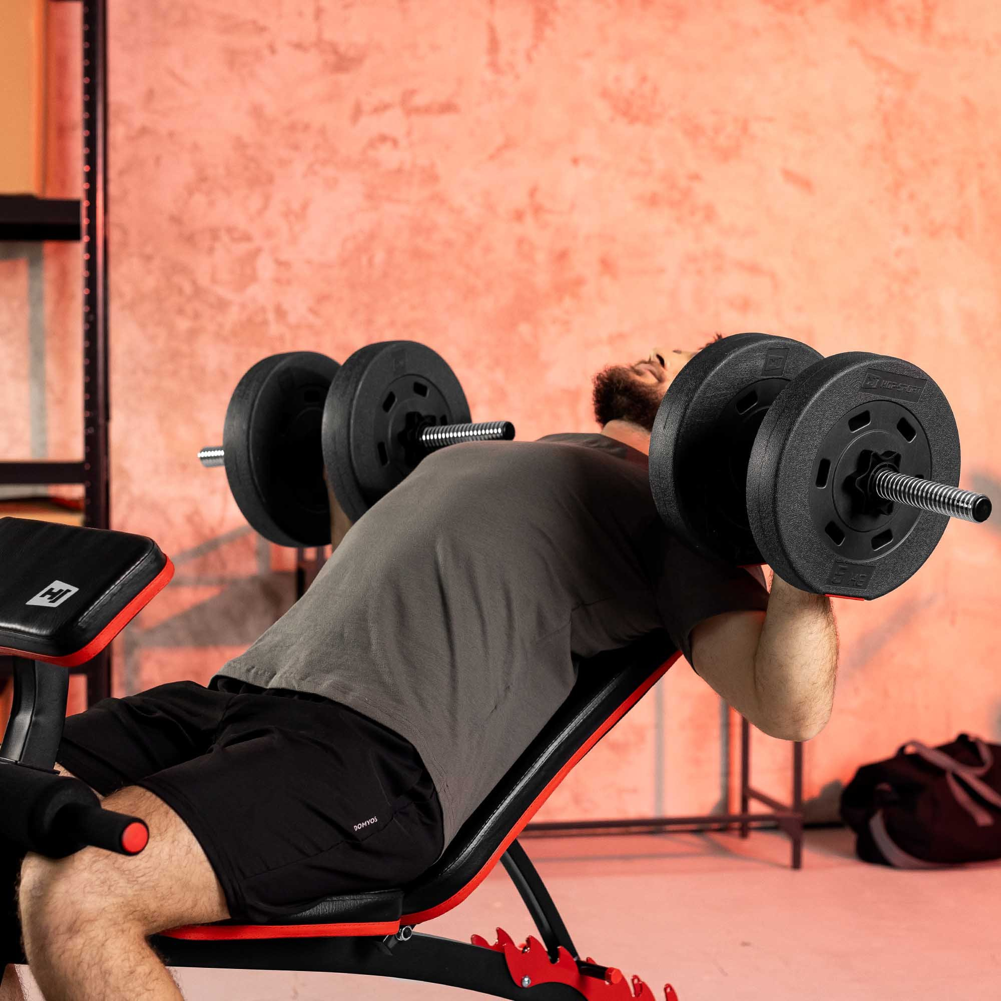 Ein Mann im grauen Shirt und schwarzen Shorts liegt auf einer Schrägbank und stemmt Hanteln im Fitnessstudio.