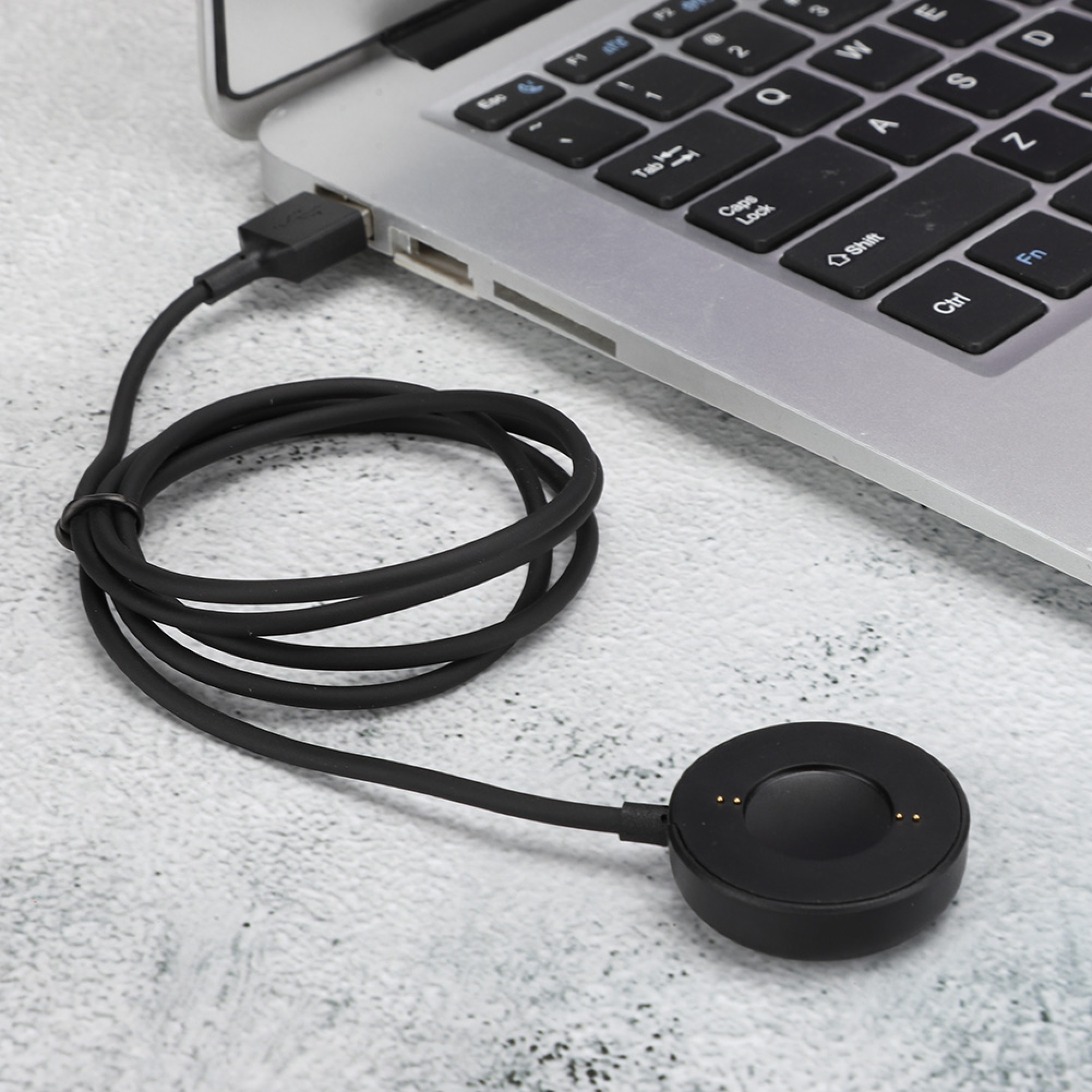 Ein schwarzes Smartwatch-Ladekabel ist an einen Laptop angeschlossen und liegt auf einer hellen Oberfläche.