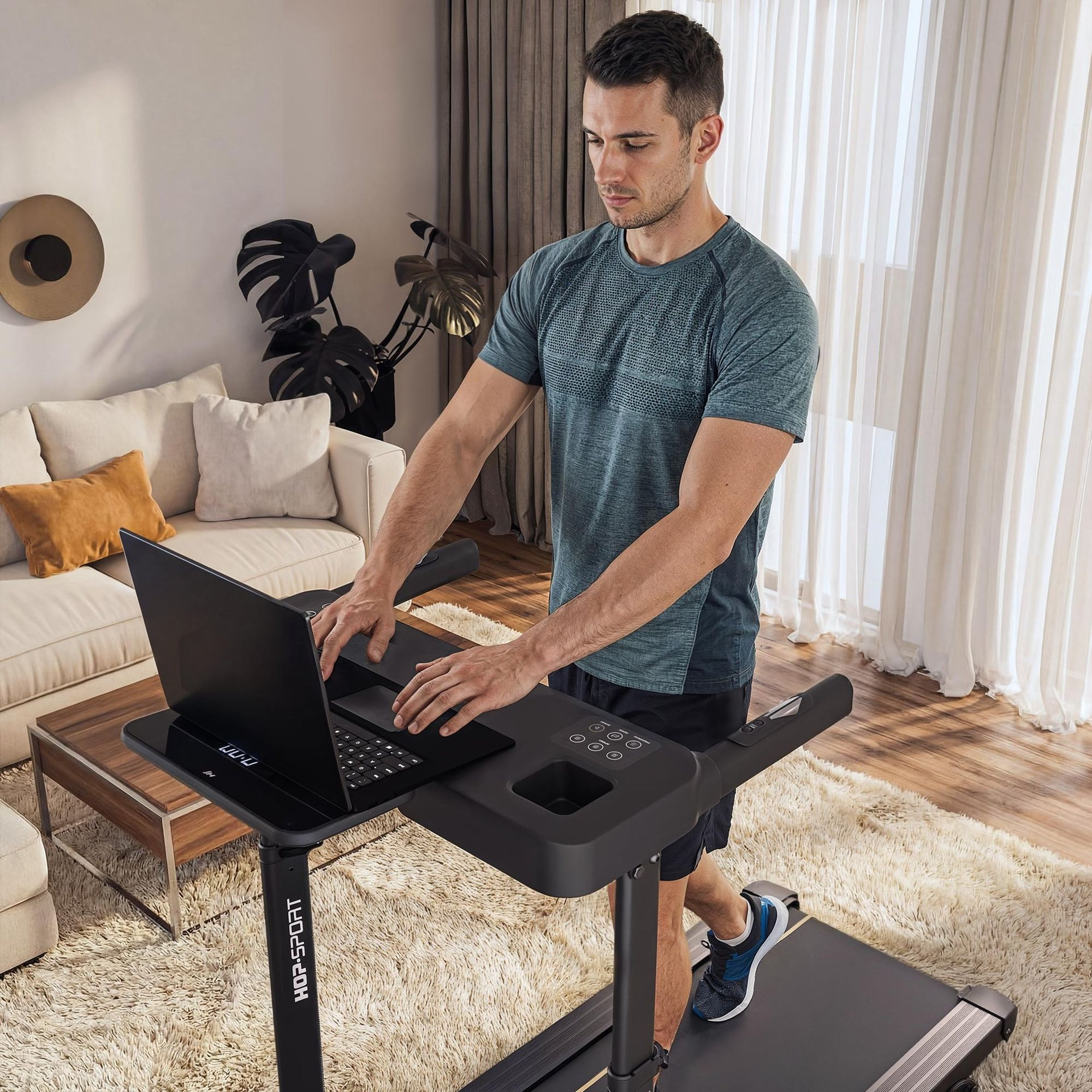 Ein Mann im blauen Shirt tippt auf einem Laptop, während er in einem Wohnzimmer auf einem Laufband geht.