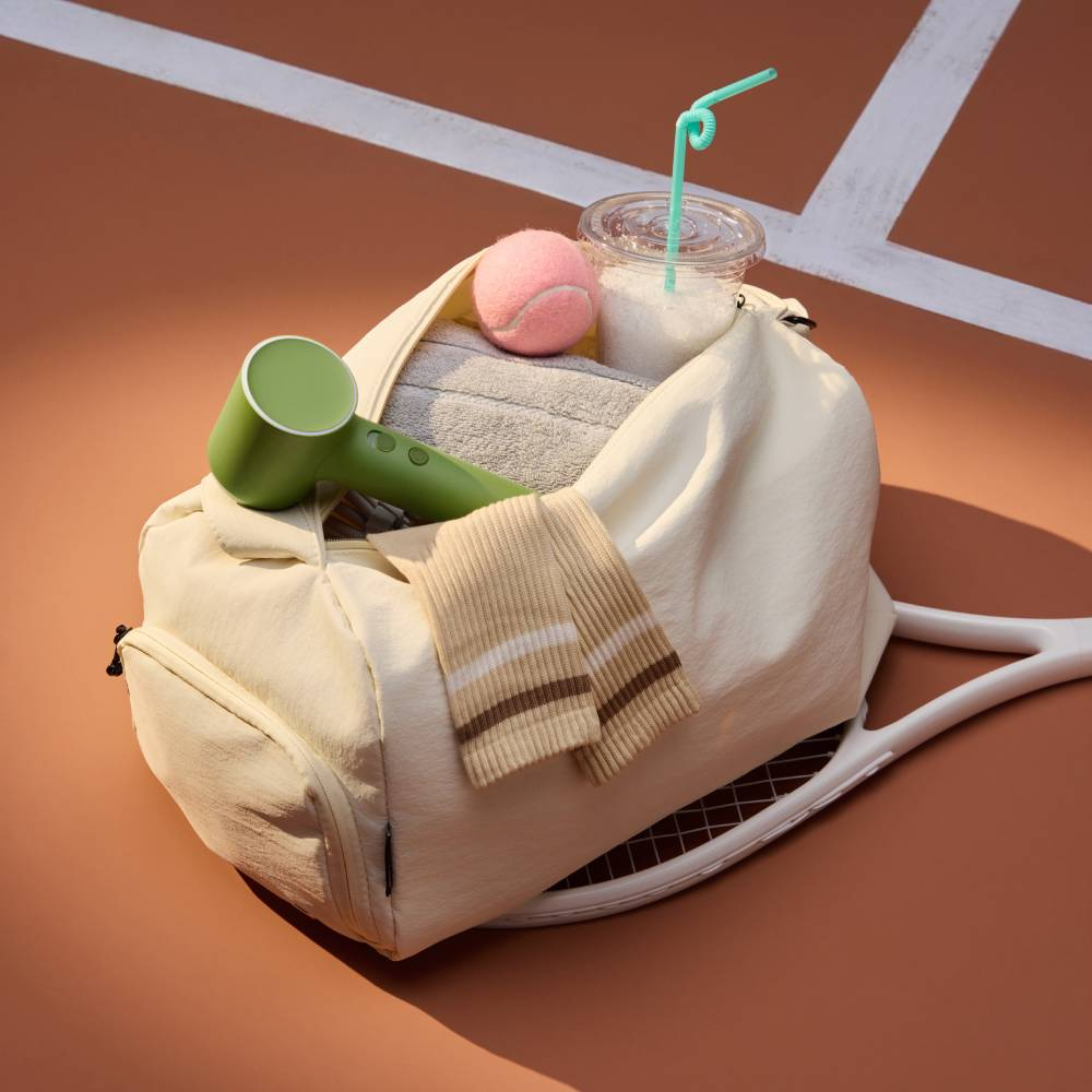 Eine weiße Sporttasche mit Tennisball, Becher, grünem Handventilator, Handtuch und Socken auf einem Tennisplatz.