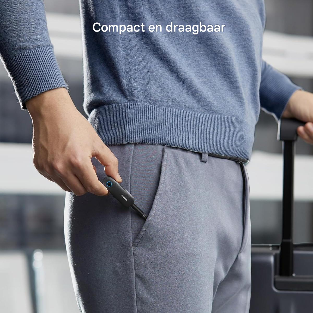Ein Mann im blauen Pullover steckt ein kleines schwarzes Gerät in seine Hose, mit einem Koffer im Hintergrund.