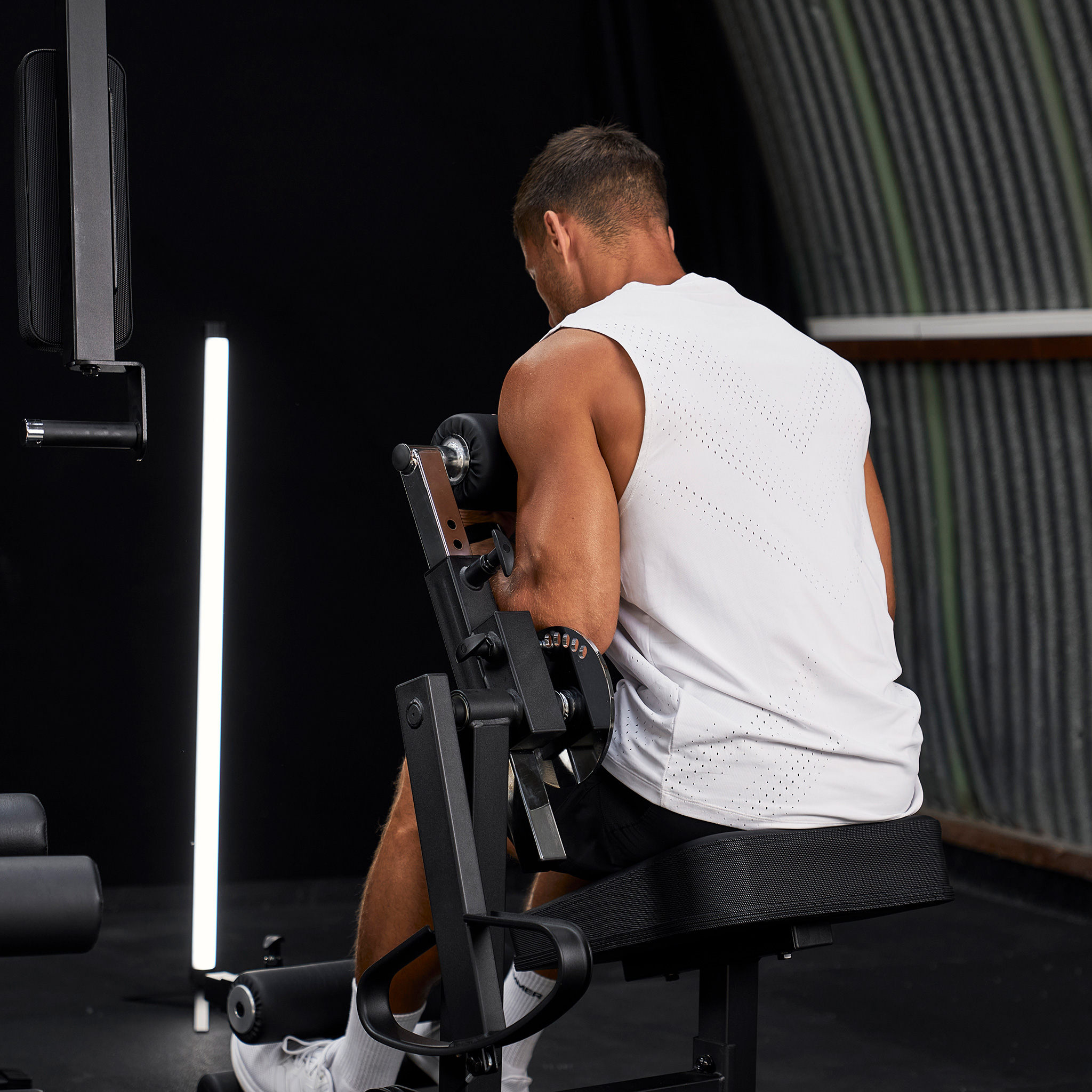 Ein Mann im weißen Tanktop sitzt in einem Fitnessstudio auf einer Scott-Curl-Maschine und trainiert.