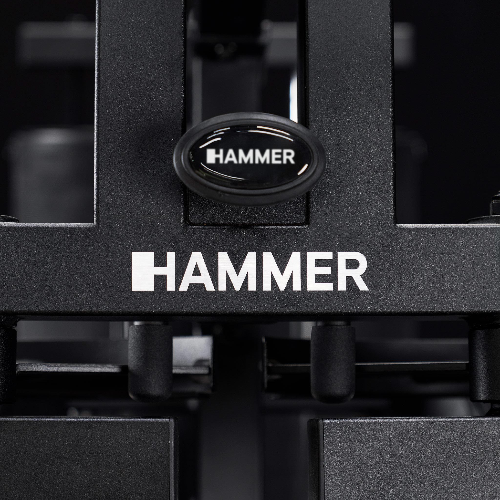 Schwarzes Hammer-Fitnessgerät mit weißem Logo und ovalem Emblem auf dunkelgrauem Hintergrund.