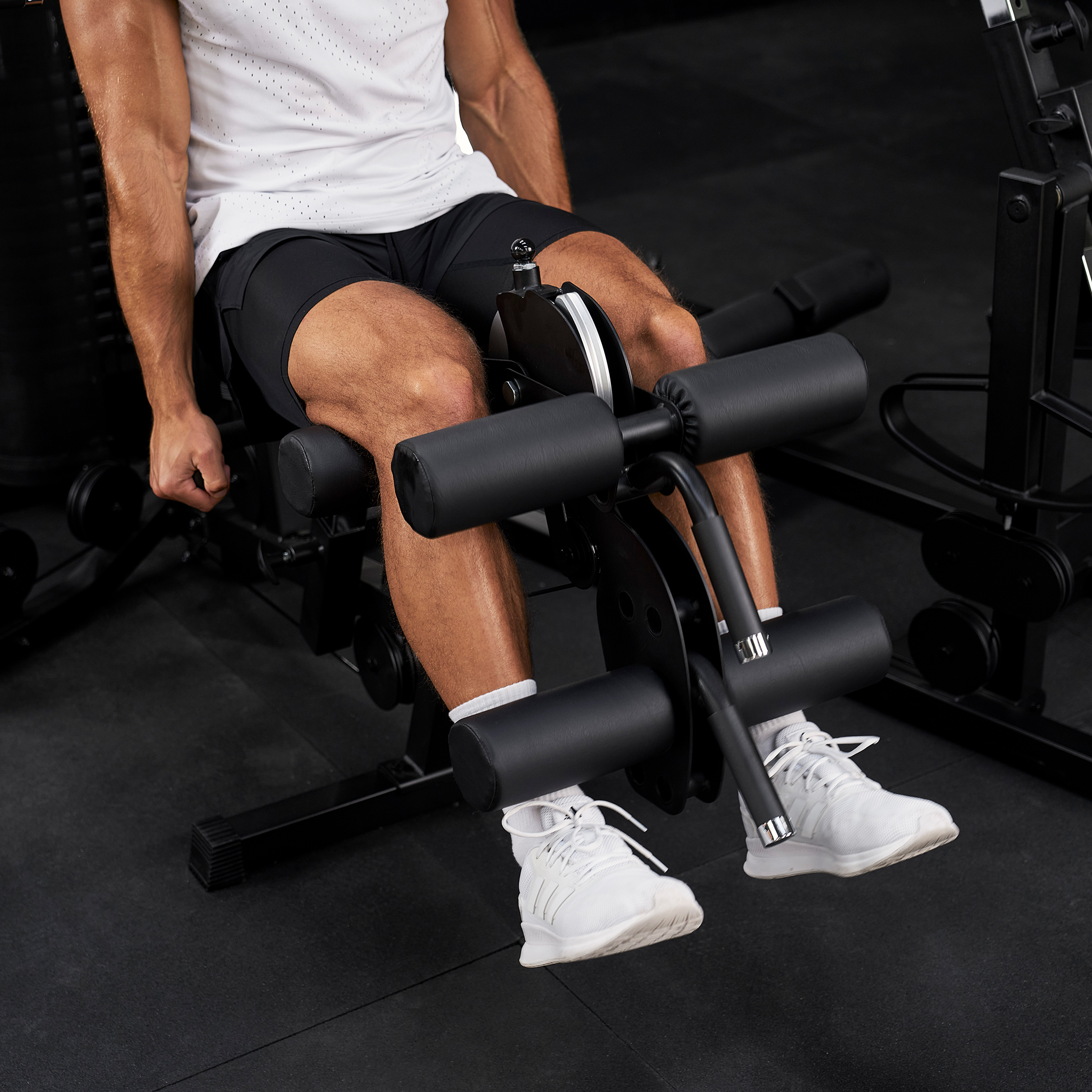 Ein Mann im Fitnessstudio trainiert an einer Beinbeugermaschine und trägt weiße Sneaker sowie schwarze Shorts.