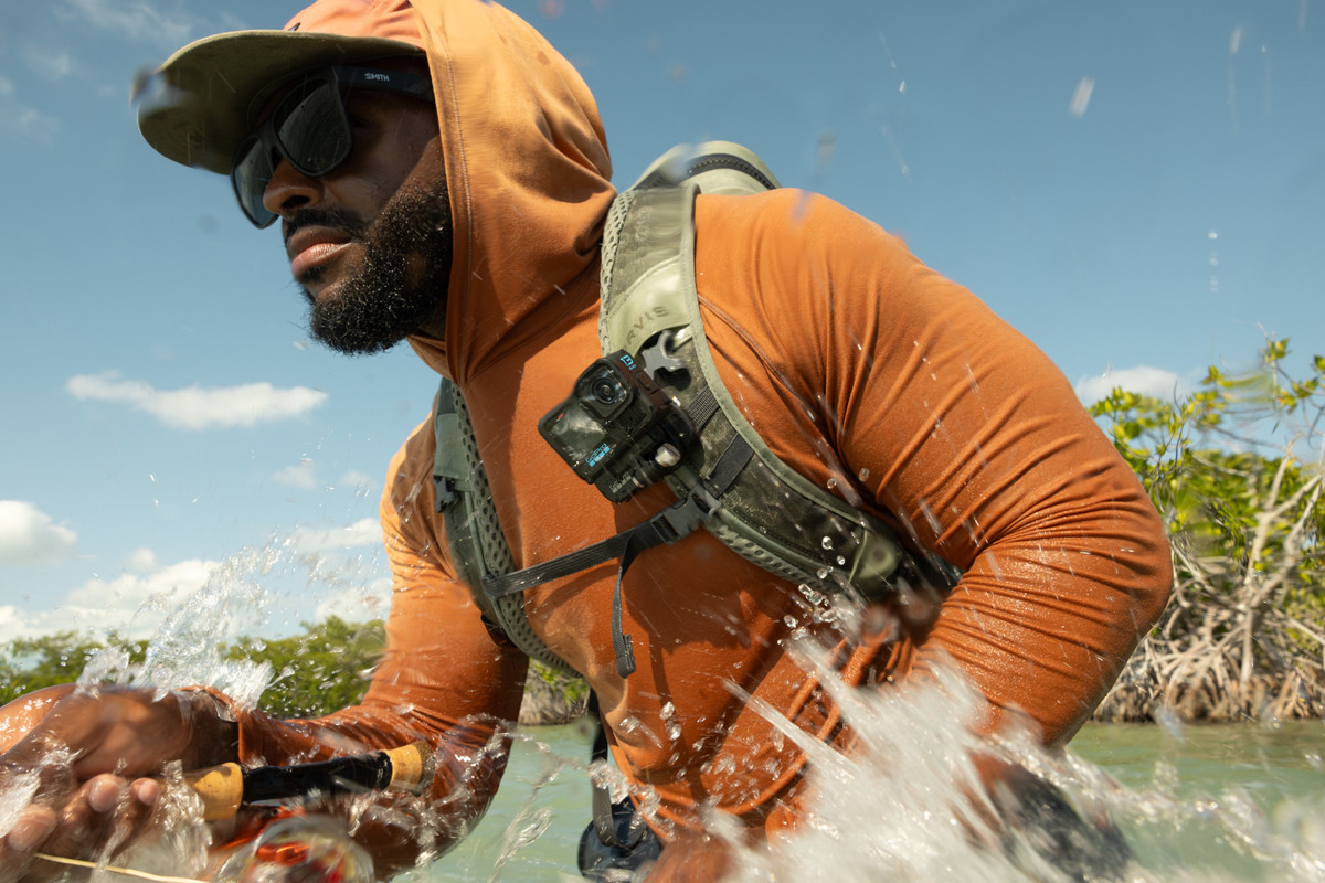 Ein Mann im orangefarbenen Hoodie und Sonnenbrille paddelt durch spritziges Wasser, Bäume und blauer Himmel im Hintergrund.
