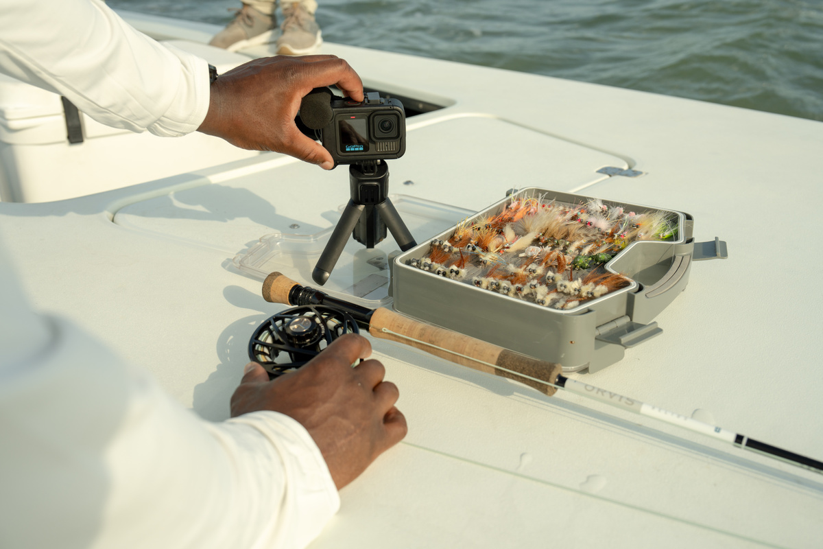 Eine Person filmt mit einer GoPro auf einem Stativ neben einer Fliegenbox und einer Angel auf einem Boot.