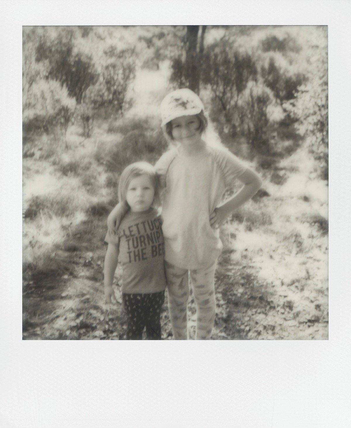 Schwarz-Weiß-Polaroid von zwei jungen Mädchen im Wald mit Tannen im Hintergrund.
