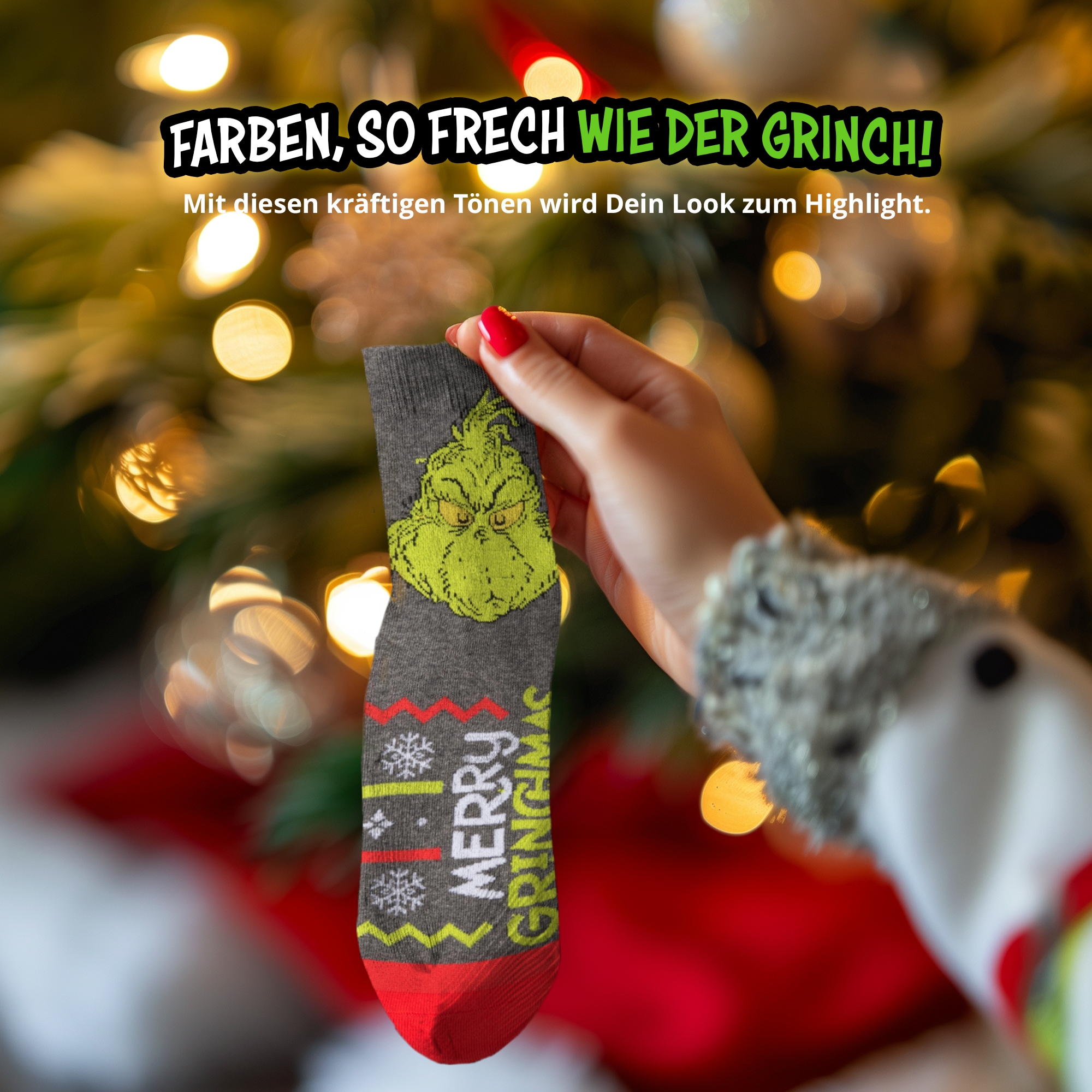 Eine Hand hält eine graue Weihnachtssocke mit Grinch-Gesicht und "Merry Grinchmas" vor einem beleuchteten Baum.