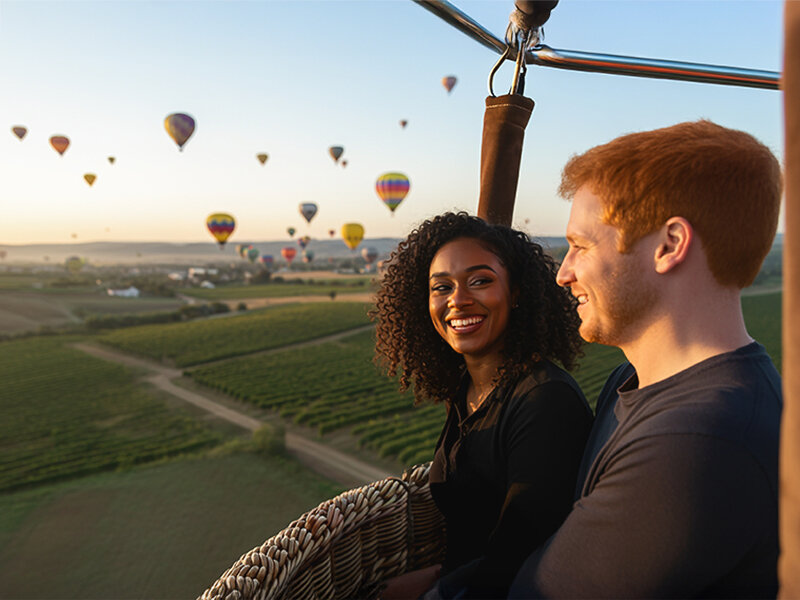 Una pareja sonriente en la cesta de un globo aerostático contempla un paisaje lleno de globos coloridos al amanecer.