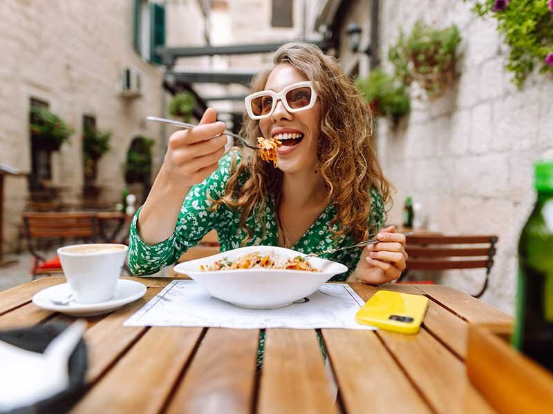 Una mujer con gafas de sol blancas y vestido verde disfruta de pasta en una mesa de madera en una calle estrecha.