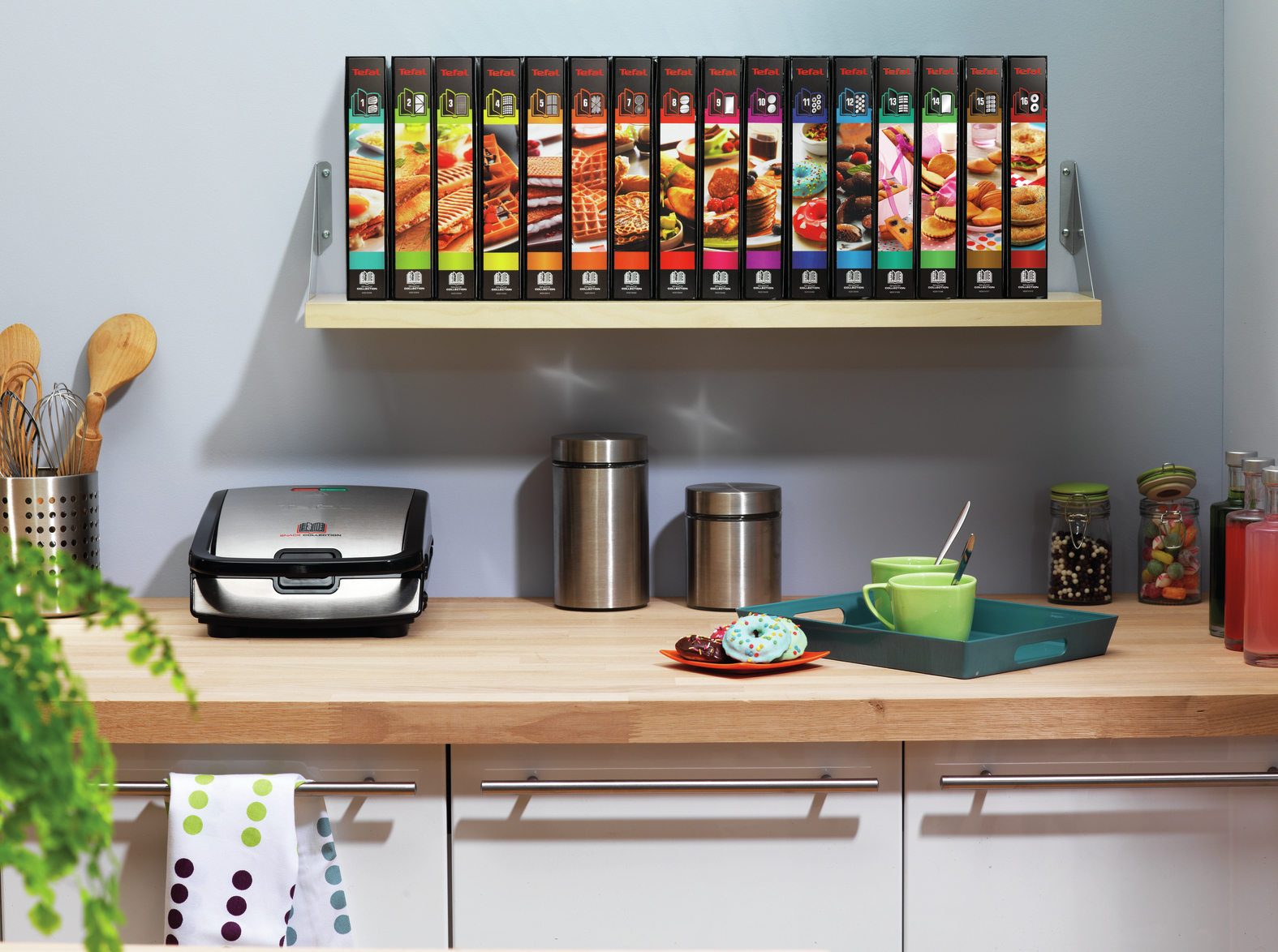 Un piano cucina con un elettrodomestico Tefal, tazze, barattoli e mensole con libri di ricette colorati.