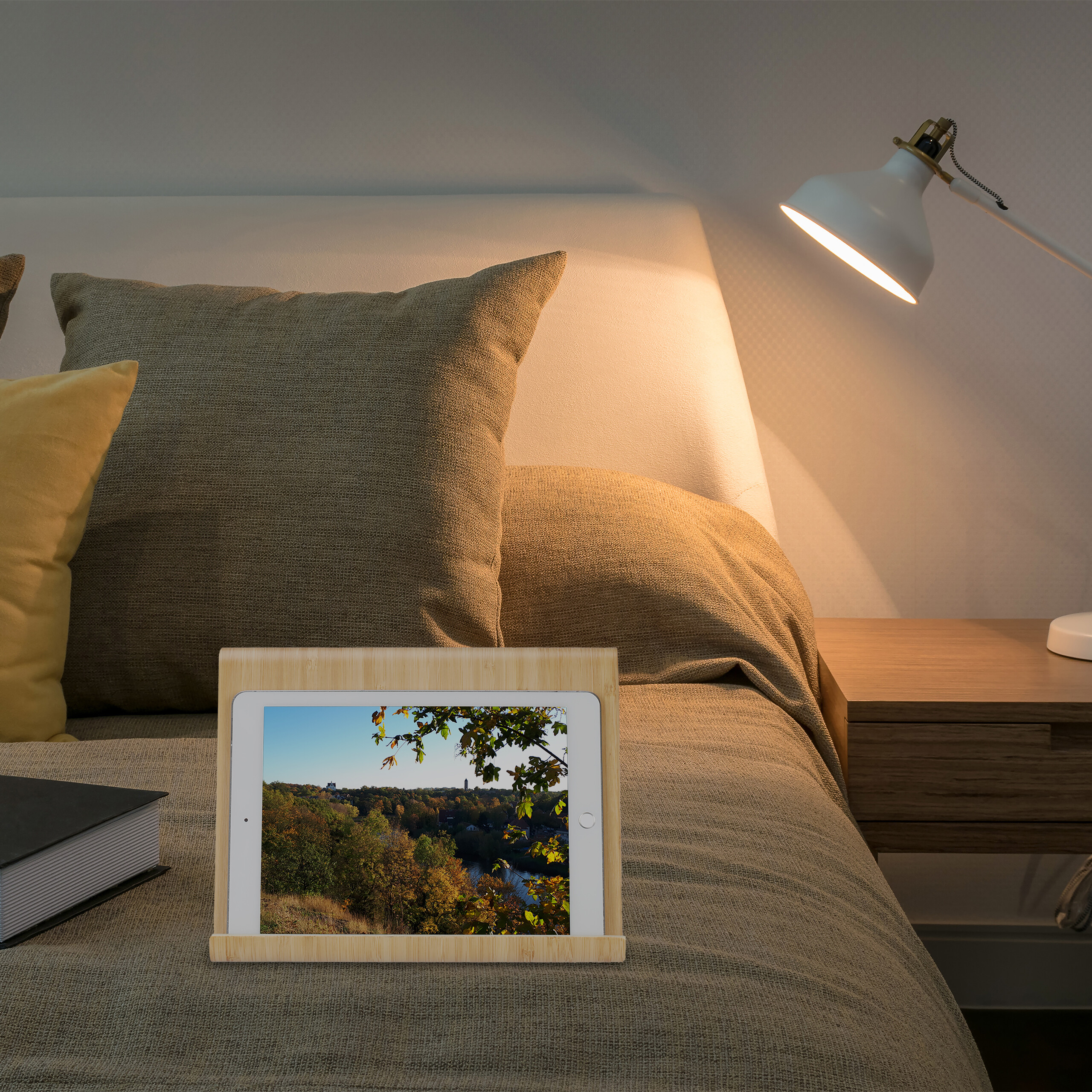 Ein Tablet in einem Holzständer auf einem Bett, daneben ein Buch und eine Lampe auf dem Nachttisch.