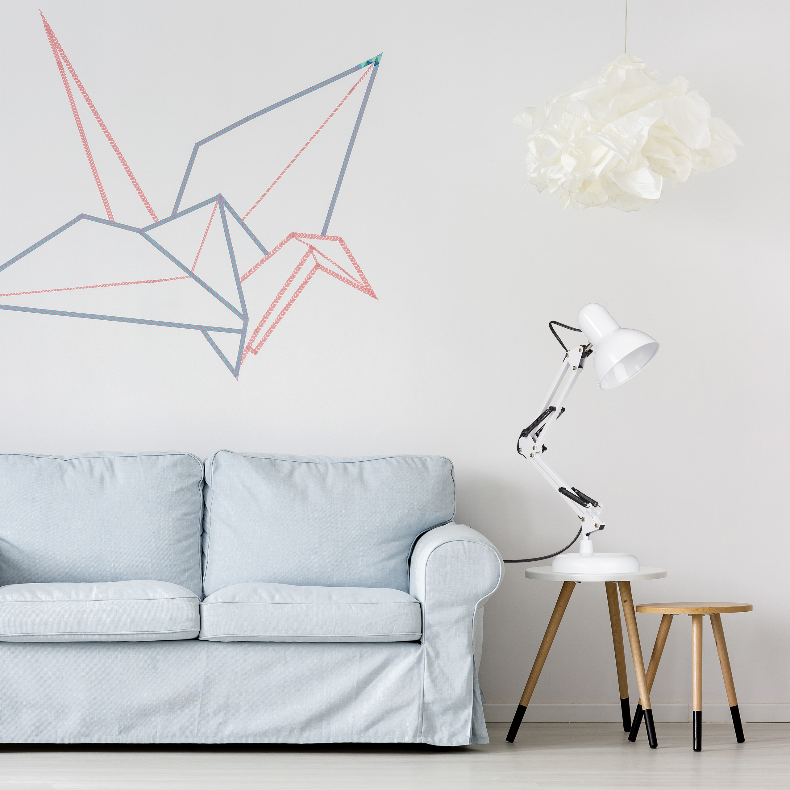 Ein hellblaues Sofa steht vor einer weißen Wand mit einem Washi-Tape-Origamivogel, Lampen und Holztischen.